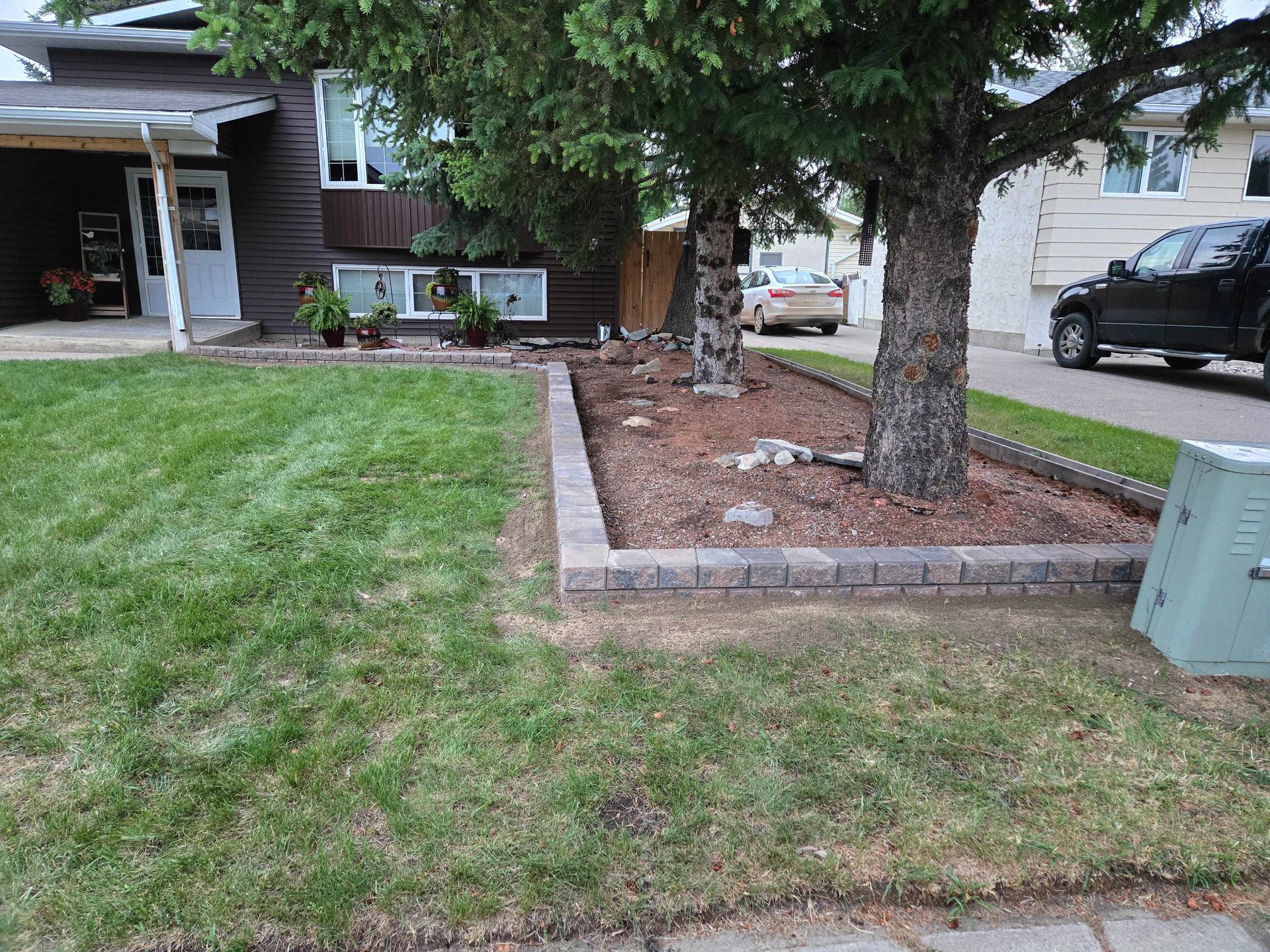 A low, rectangular brick retaining wall surrounds two large trees with mulch in a suburban front yard.