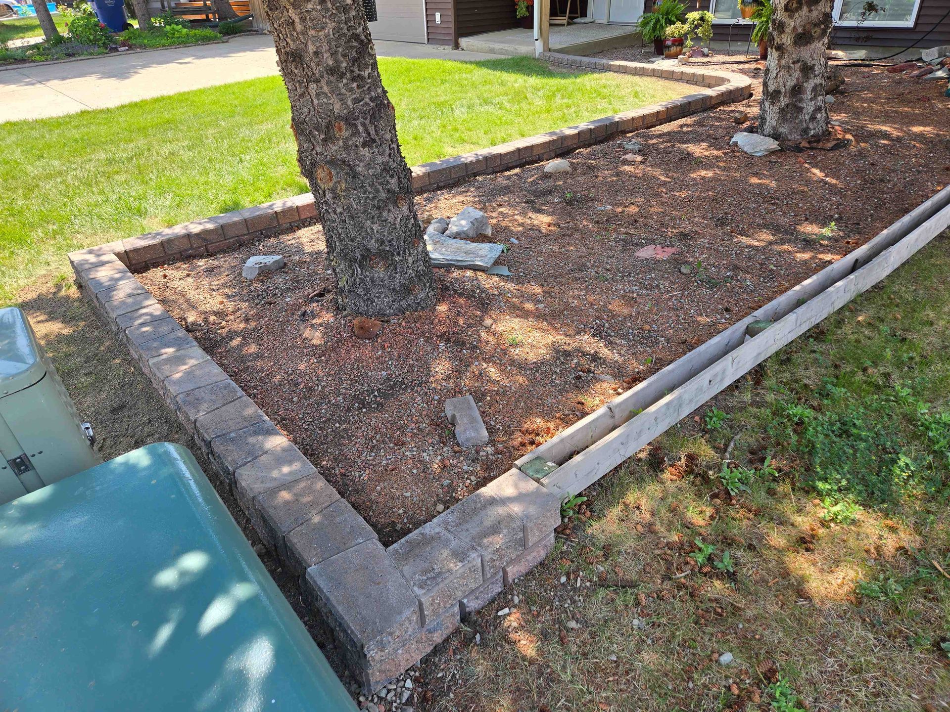 A garden bed with stone edging and wood mulch, containing two trees and scattered rocks, set next to a grassy lawn.
