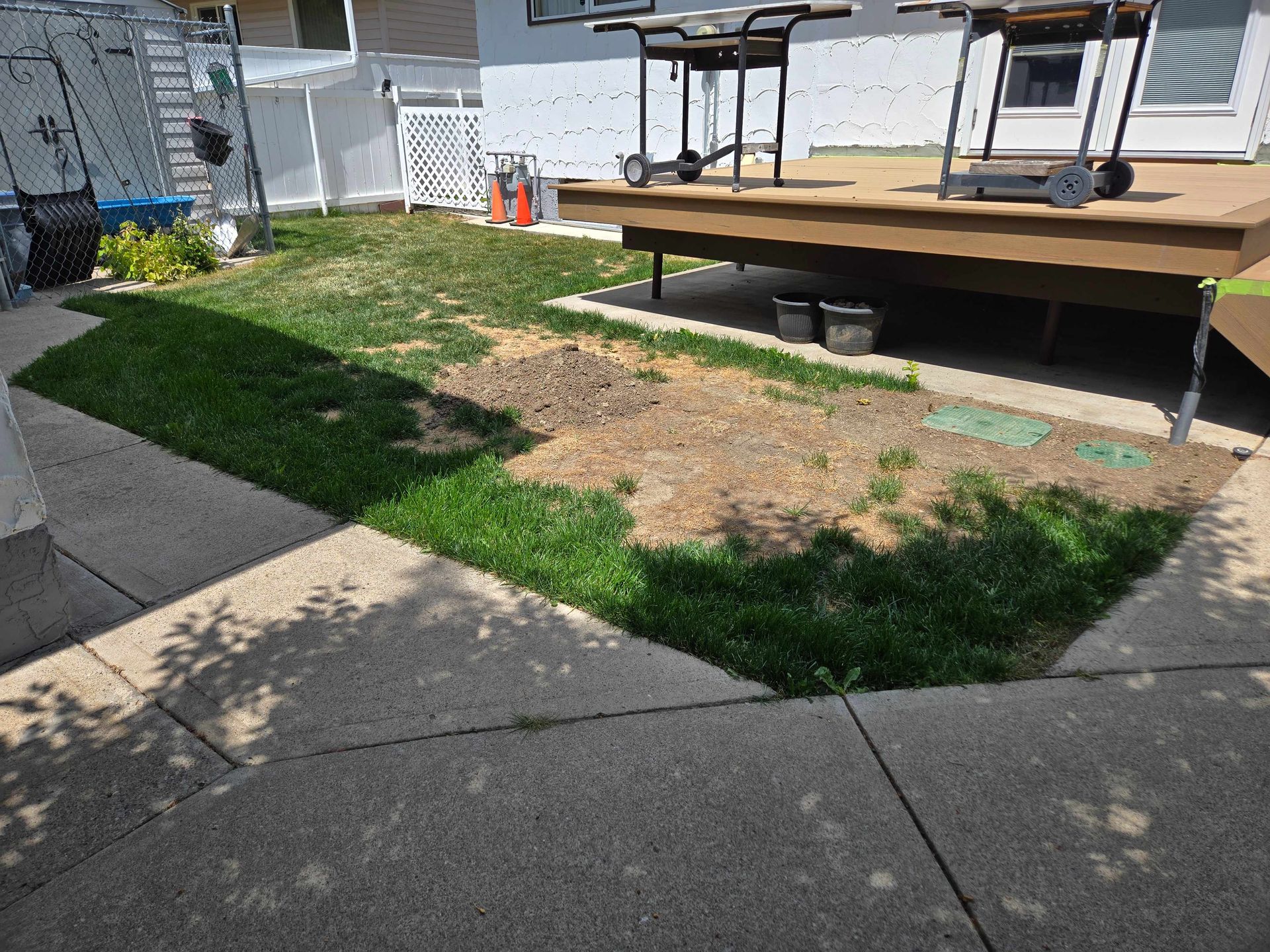 A small residential backyard with a partially patchy lawn, a wooden deck, and a concrete patio path.