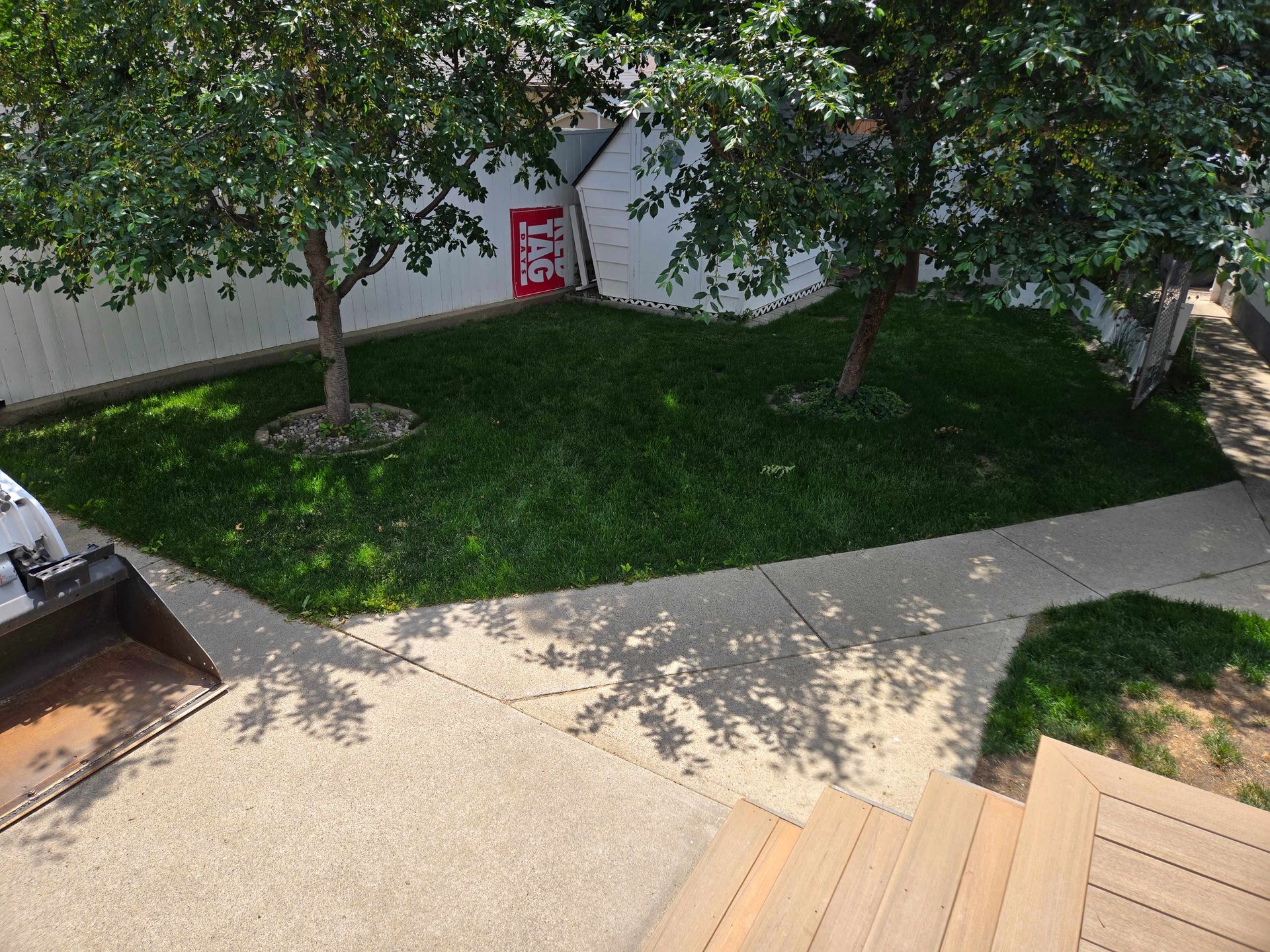 A high-angle view of a backyard with green grass, two trees, a concrete walkway, and wooden stairs in the foreground.