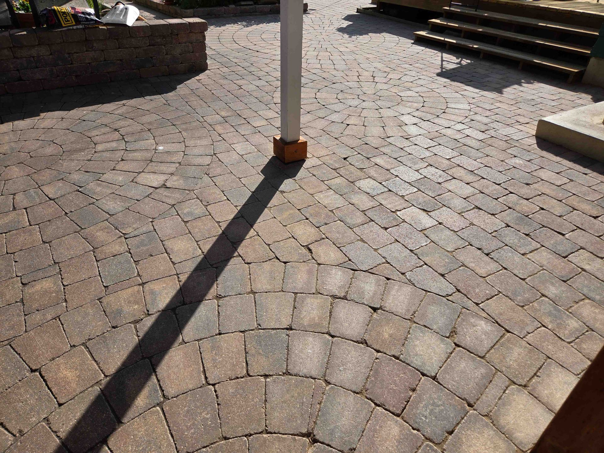 A close-up of a brown, patterned paver patio featuring circular stone layouts and a white support post casting a shadow.