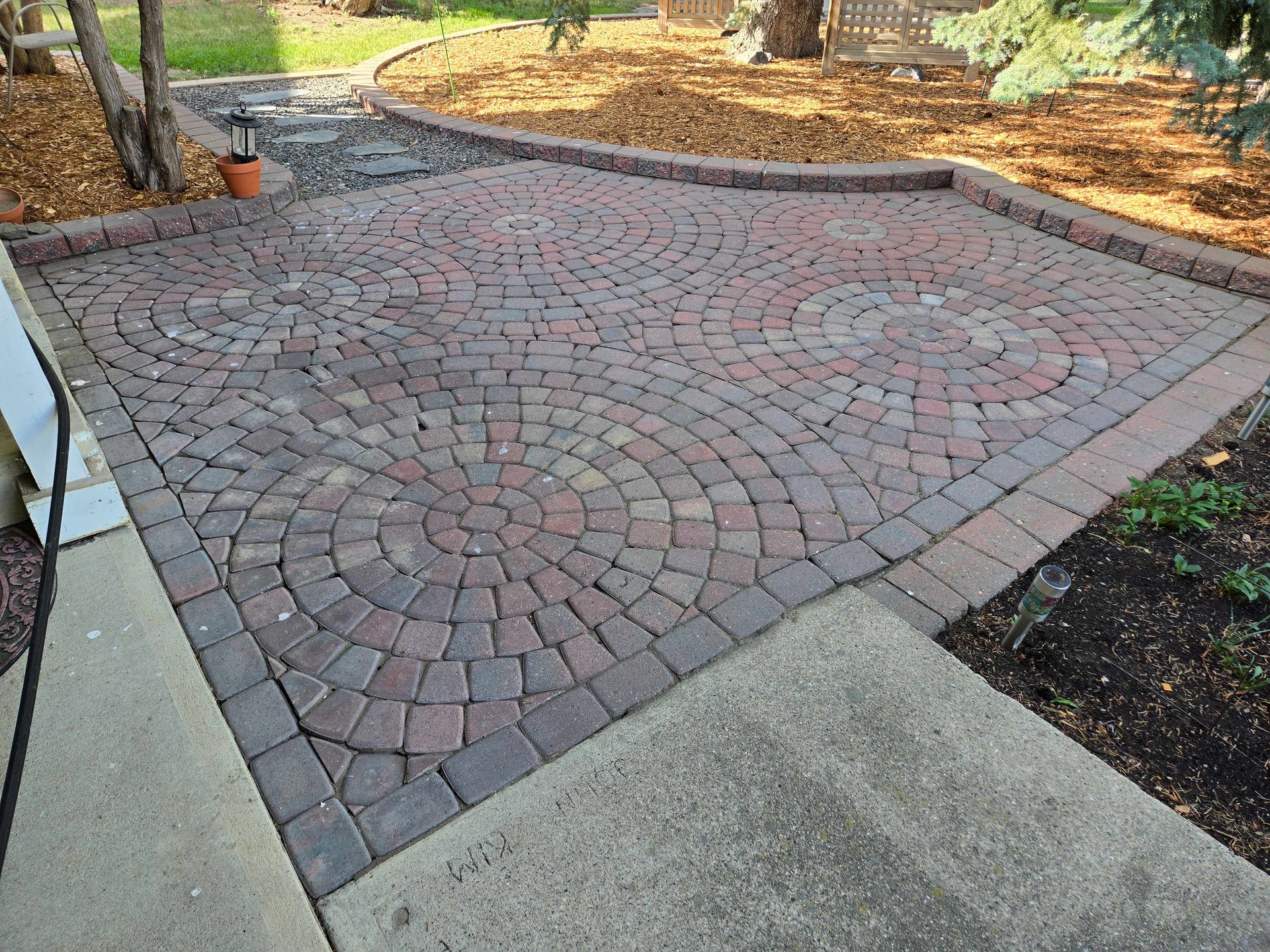 A patio featuring circular, fan-patterned red and brown pavers bordered by a darker masonry edge in a backyard garden.
