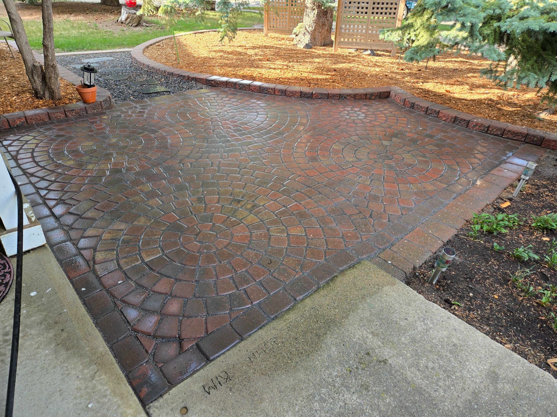 A patio made of circular-patterned, reddish-brown bricks, surrounded by mulch, trees, and a concrete walkway.