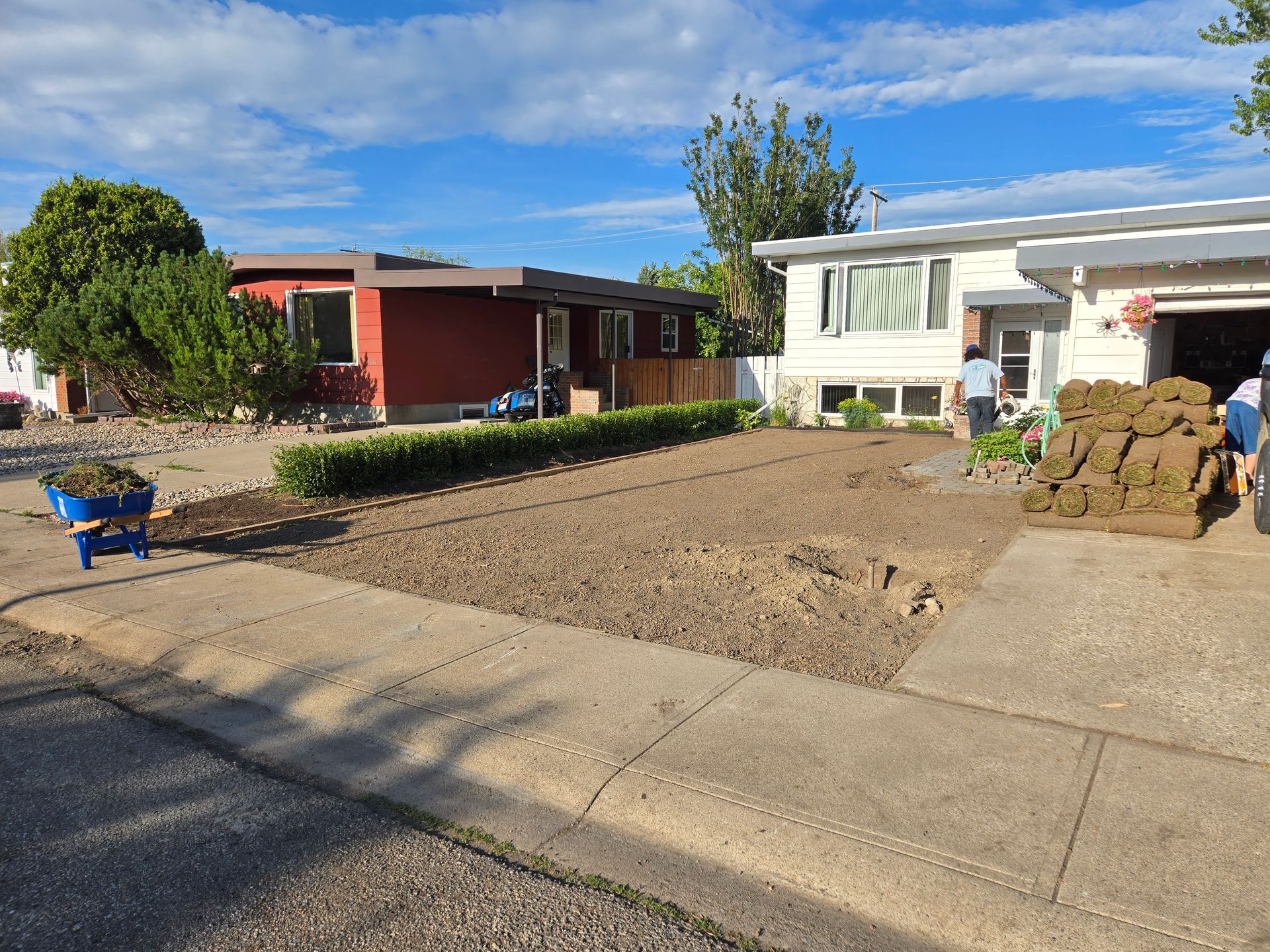 Front yard under construction beside two homes, with dirt, stacked materials, and a worker near the driveway.