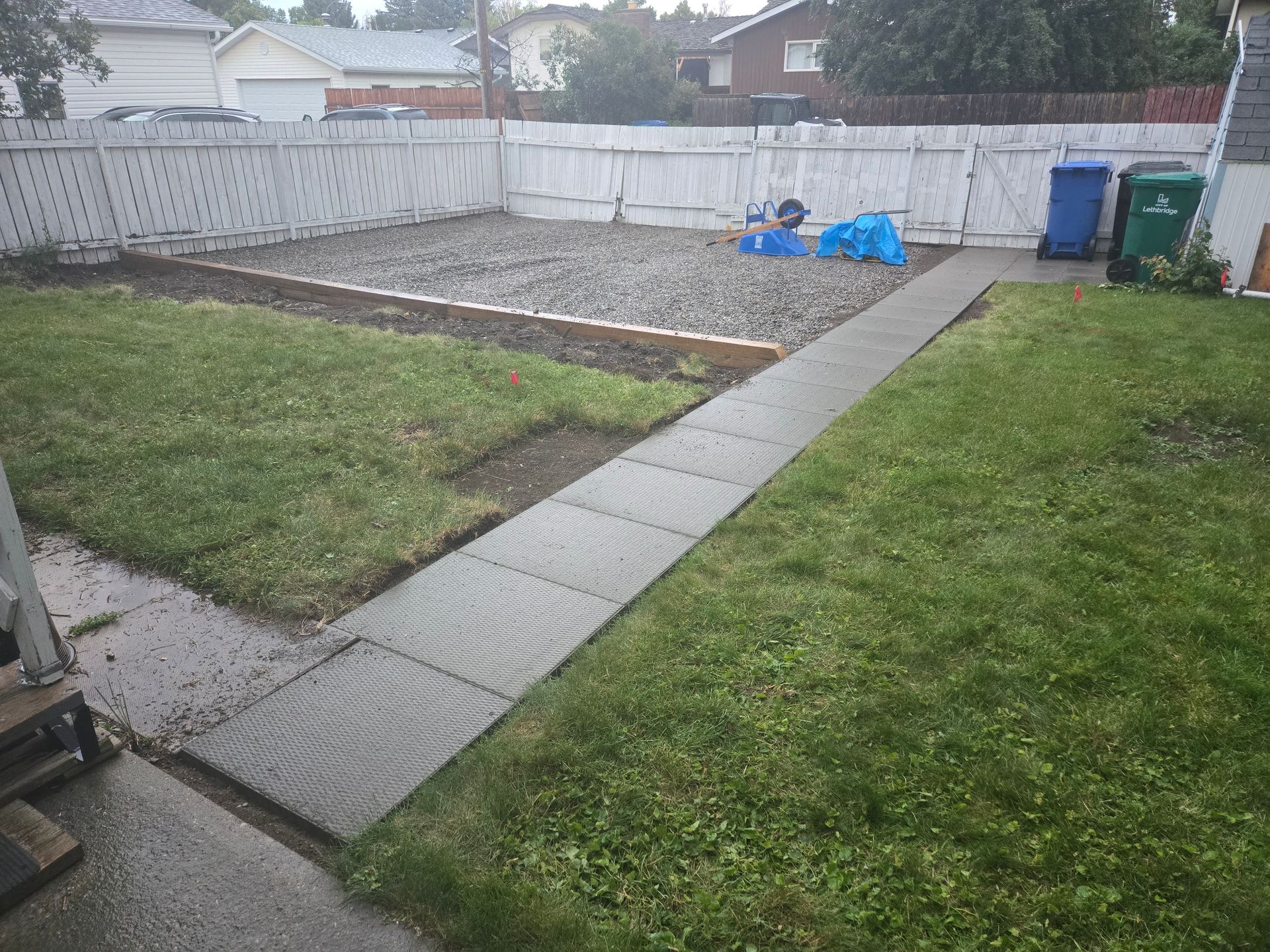 Backyard with a concrete path, grass, white fence, and blue lawn chairs near trash bins