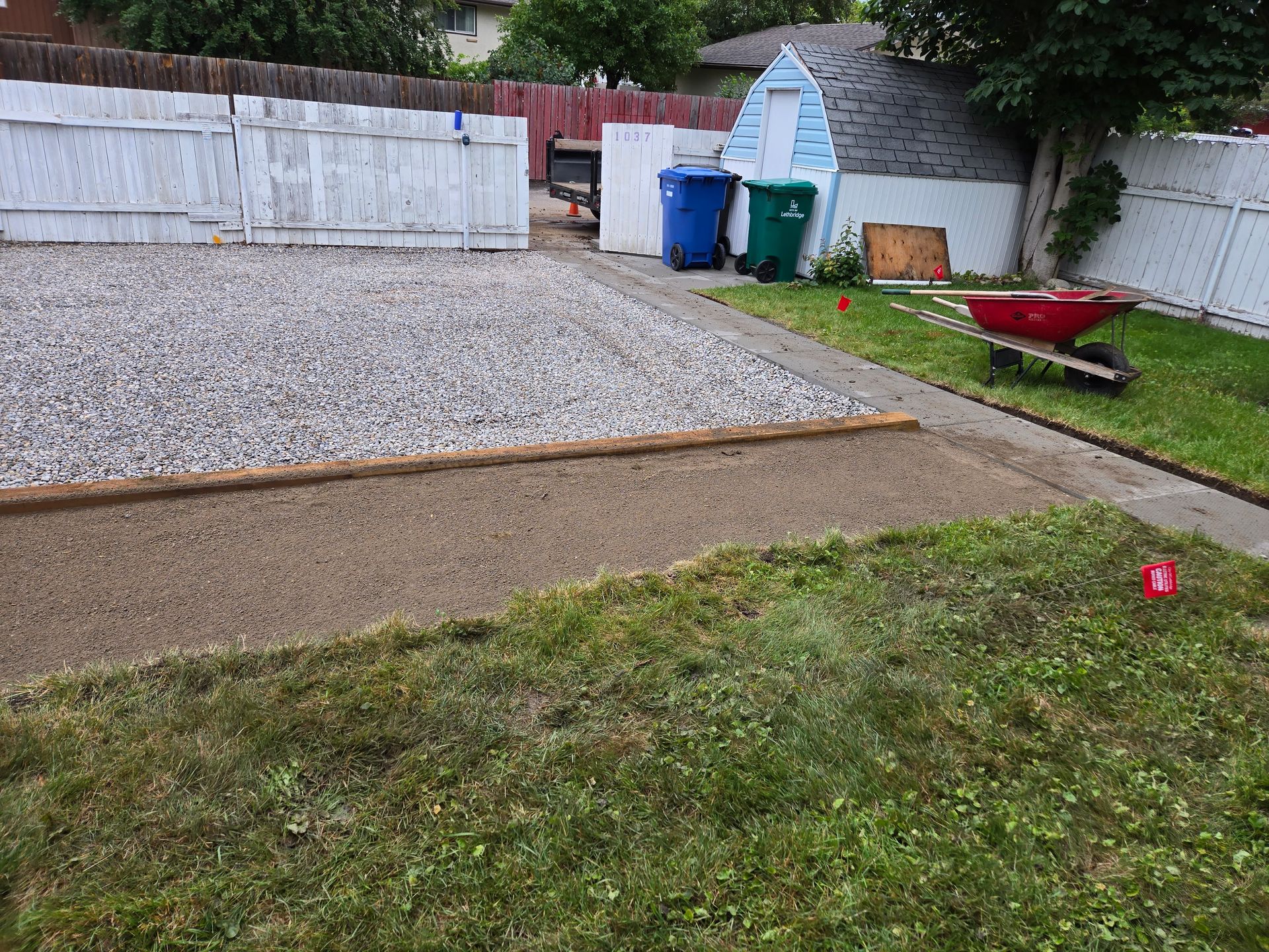 Backyard with gravel patio, grass, white fence, blue bins, and a red wheelbarrow near the wall