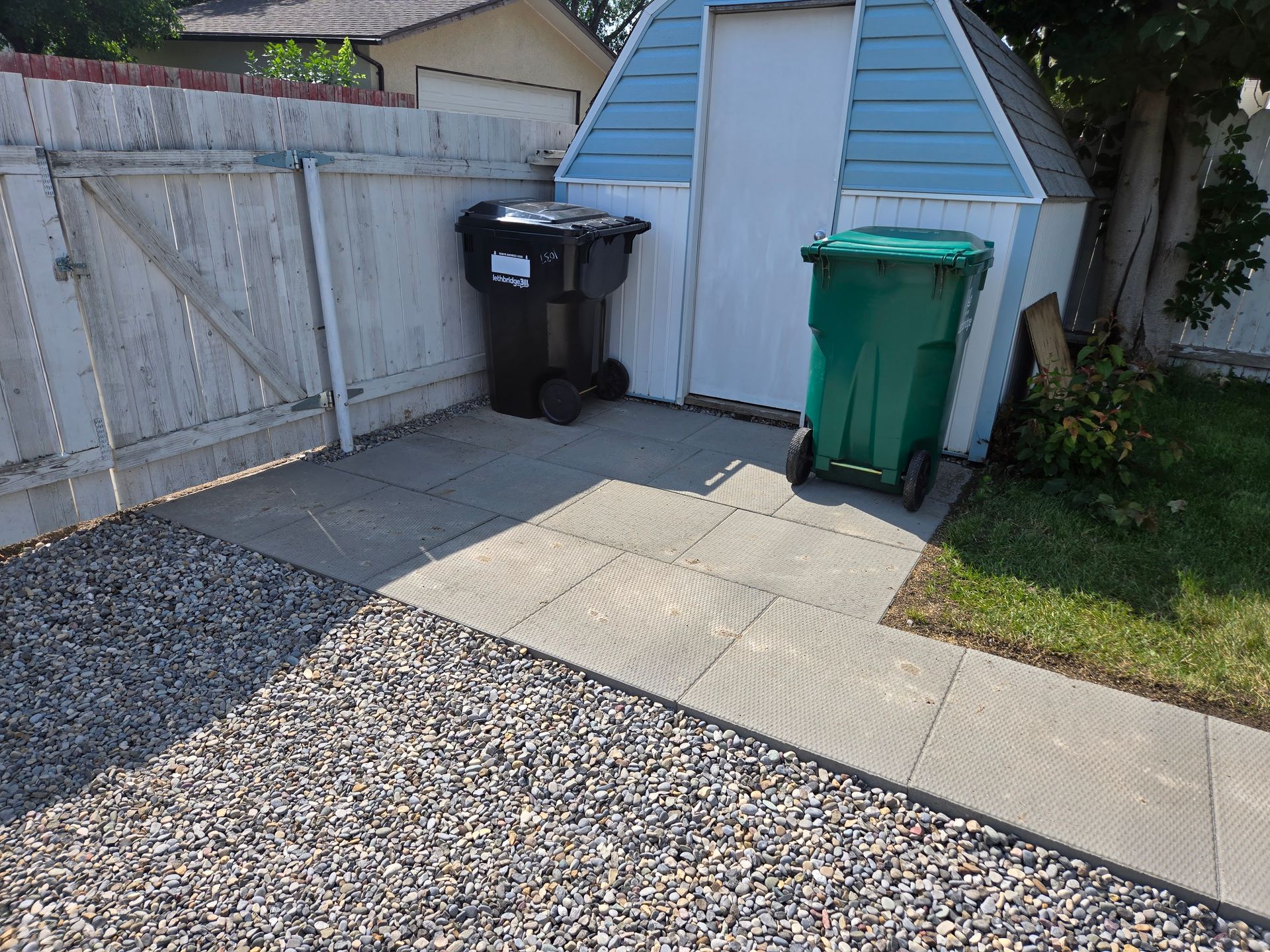 Backyard shed beside a wooden fence, with black and green trash bins on a paved patio.