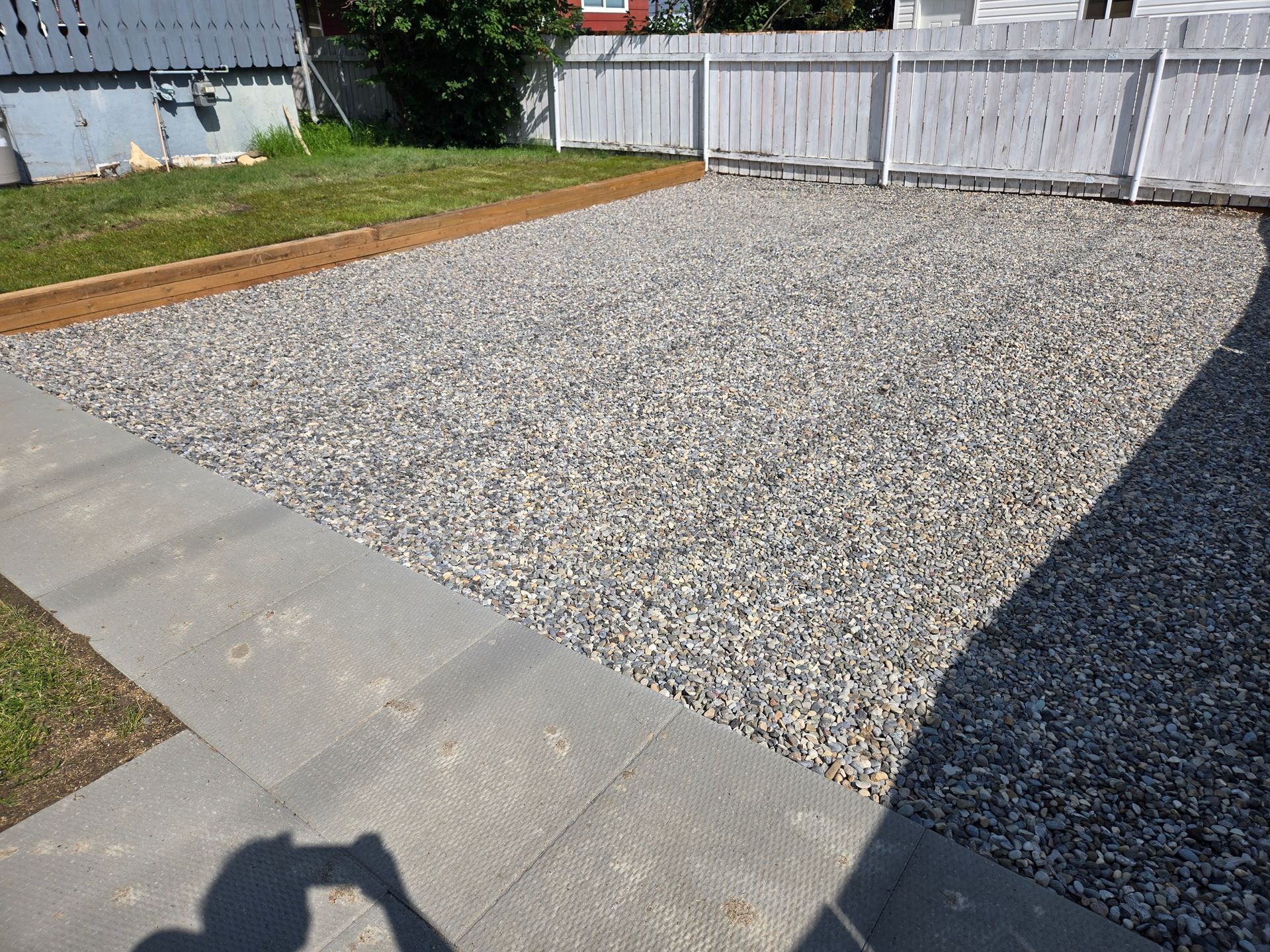 Gravel backyard with concrete border, white fence, and grass along the edges under bright sun