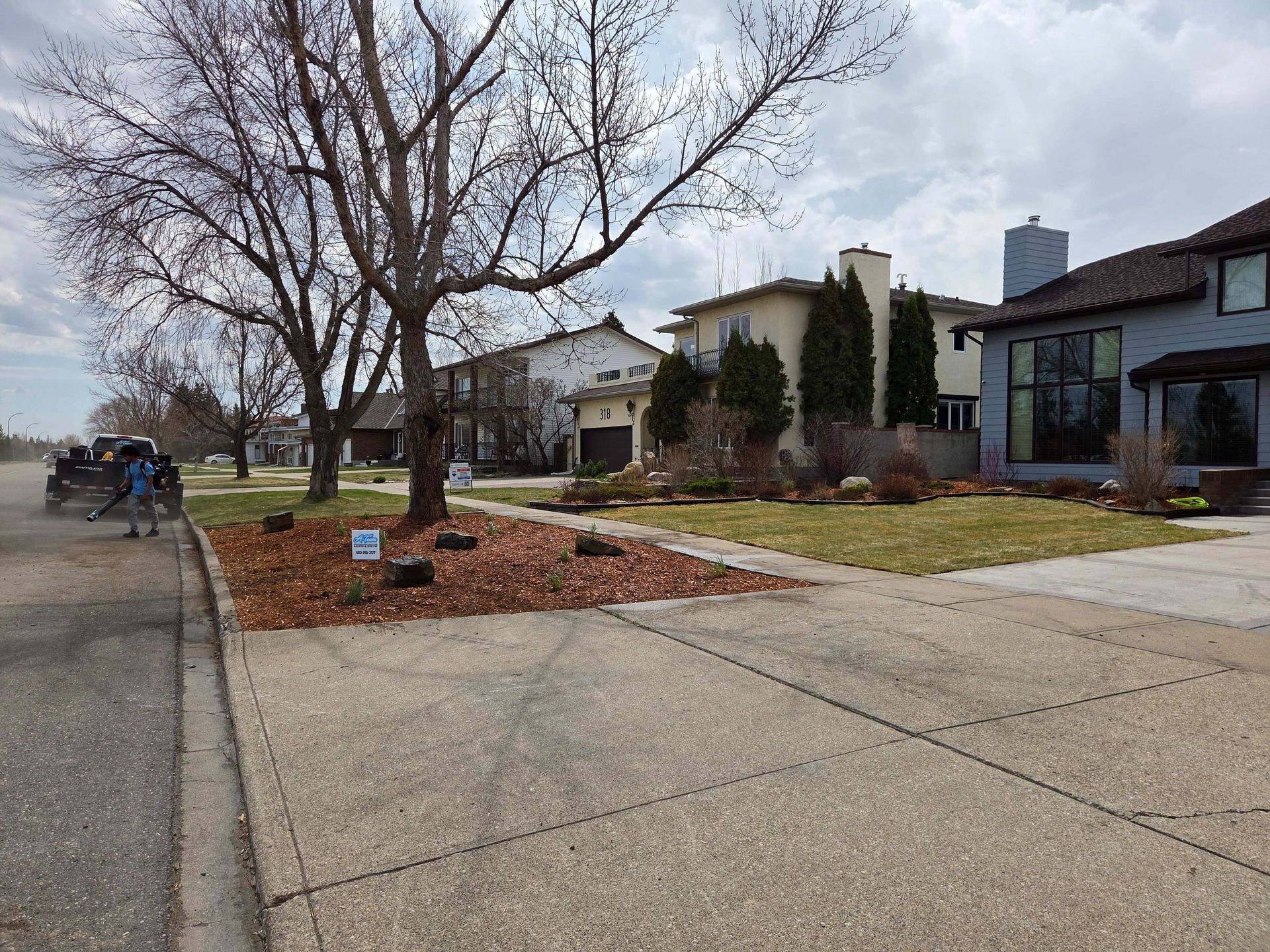 Residential street with bare trees, brick landscaping, and modern houses on a cloudy day