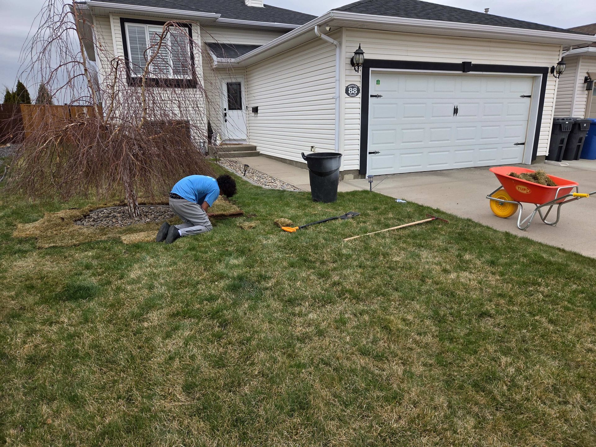 Person kneeling in a front yard with a tool near a house, beside a wheelbarrow and garage.