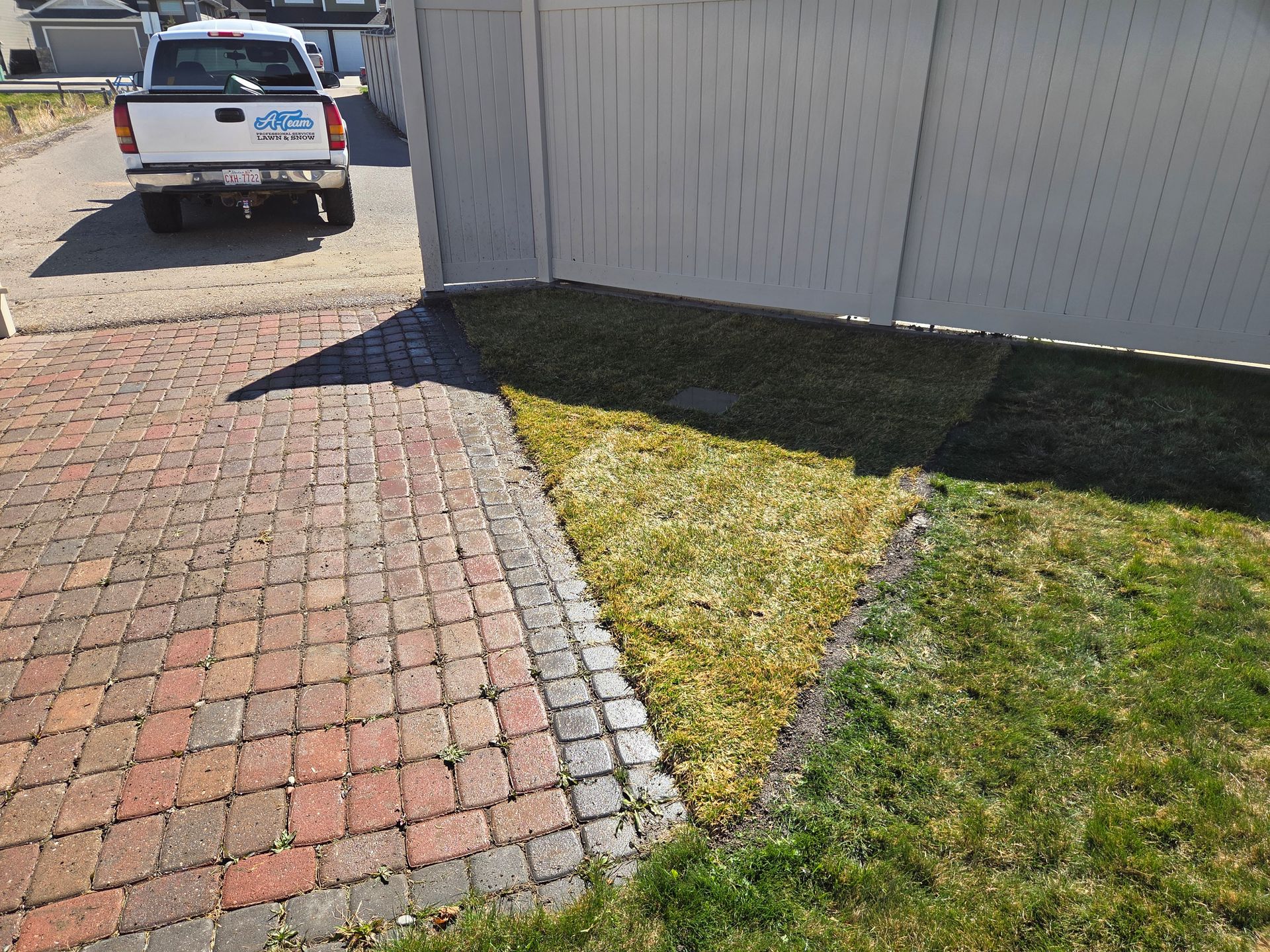 Brick driveway beside a white house, with a patch of grass and a parked truck in the background