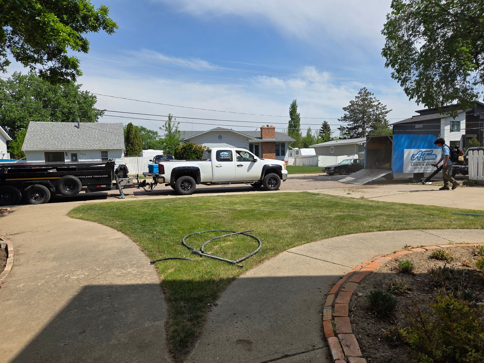 Truck and white van parked on a driveway beside a grassy yard and blue outbuilding on a sunny day
