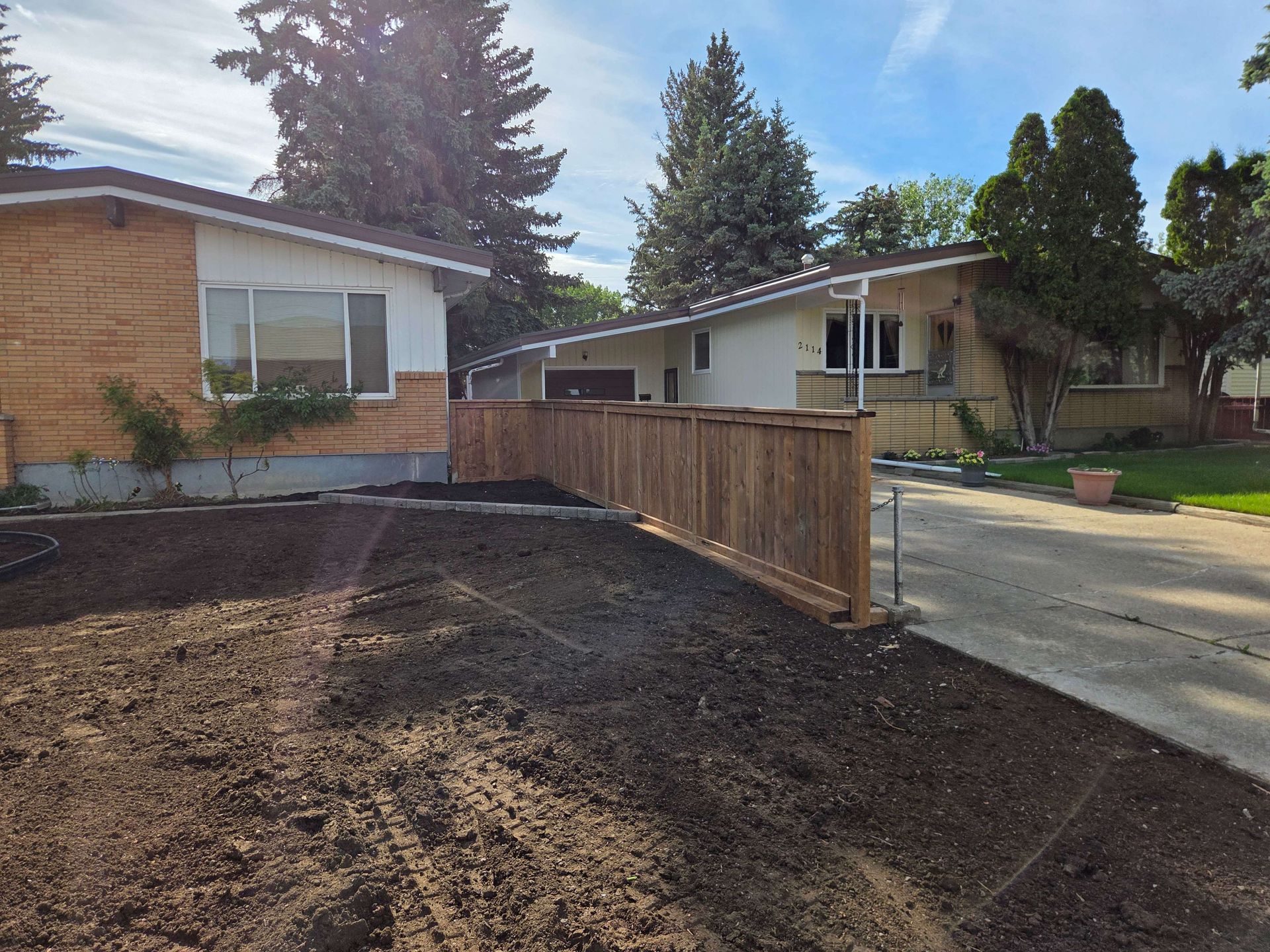 Residential street with a wooden fence, dirt yard, and brick houses under a blue sky