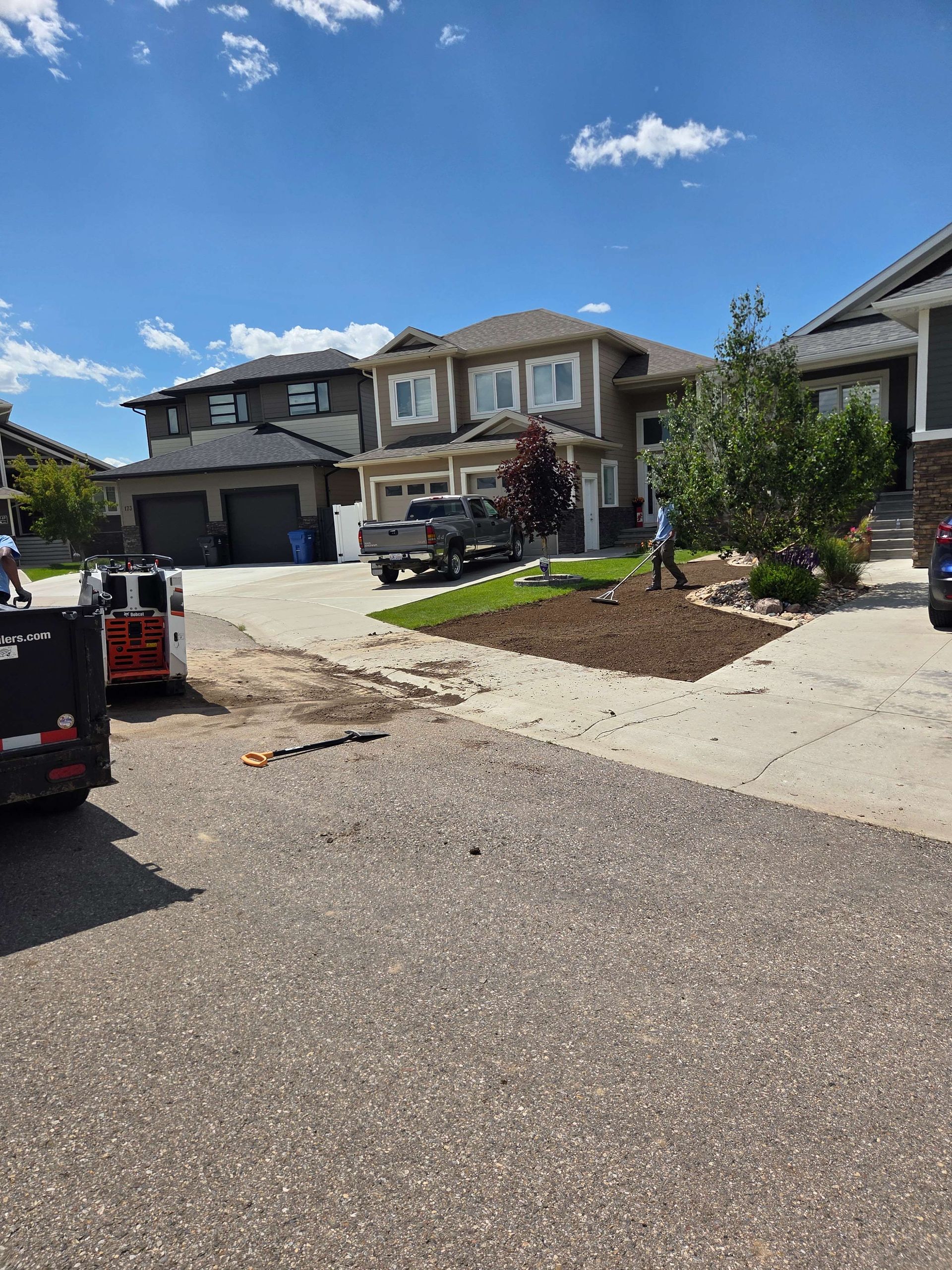 Suburban street with new homes, gravel driveway, and workers landscaping under a bright blue sky