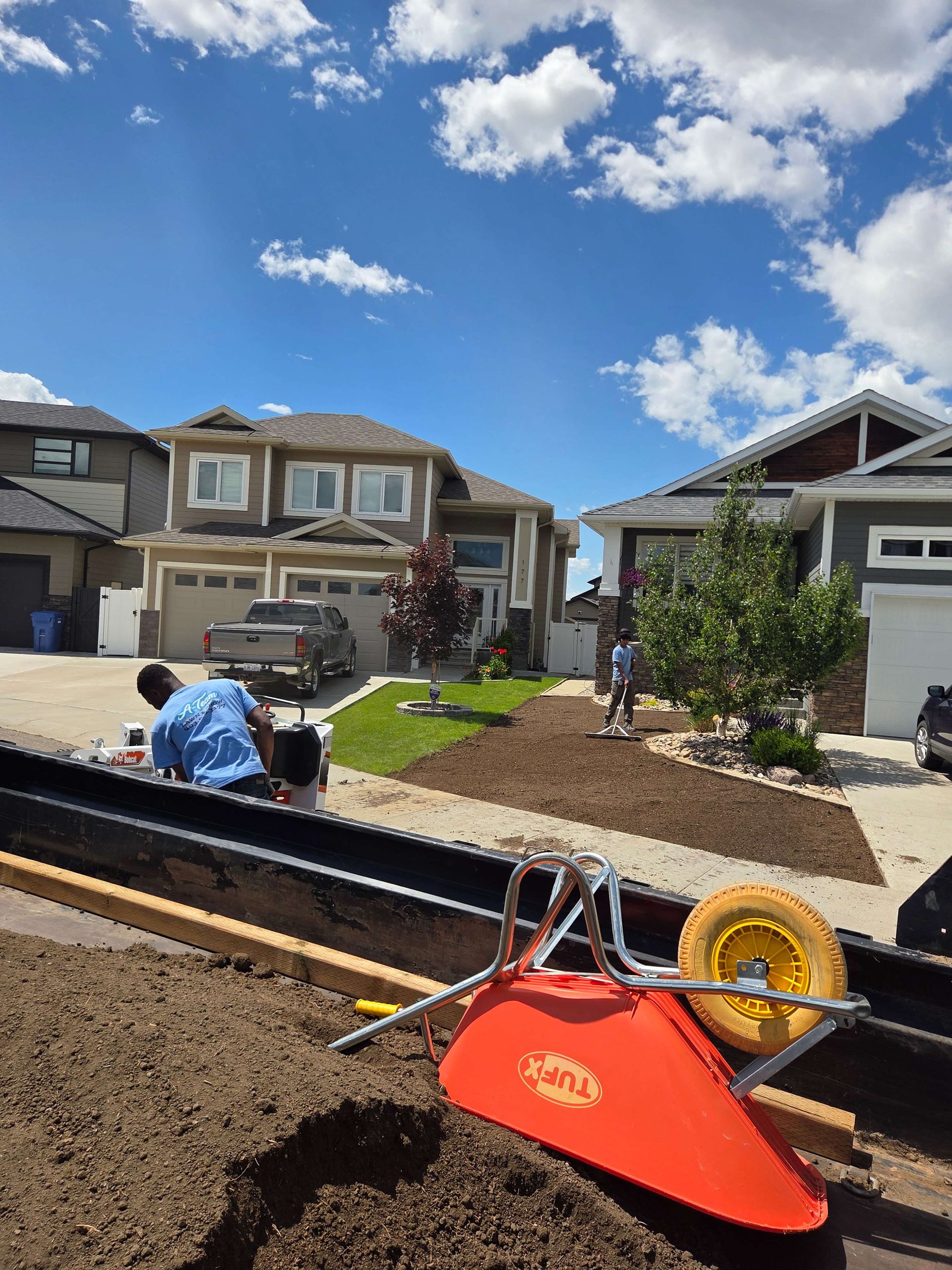 Roadwork in a suburban neighborhood, with orange wheelbarrow and gravel pile in the foreground under a blue sky.