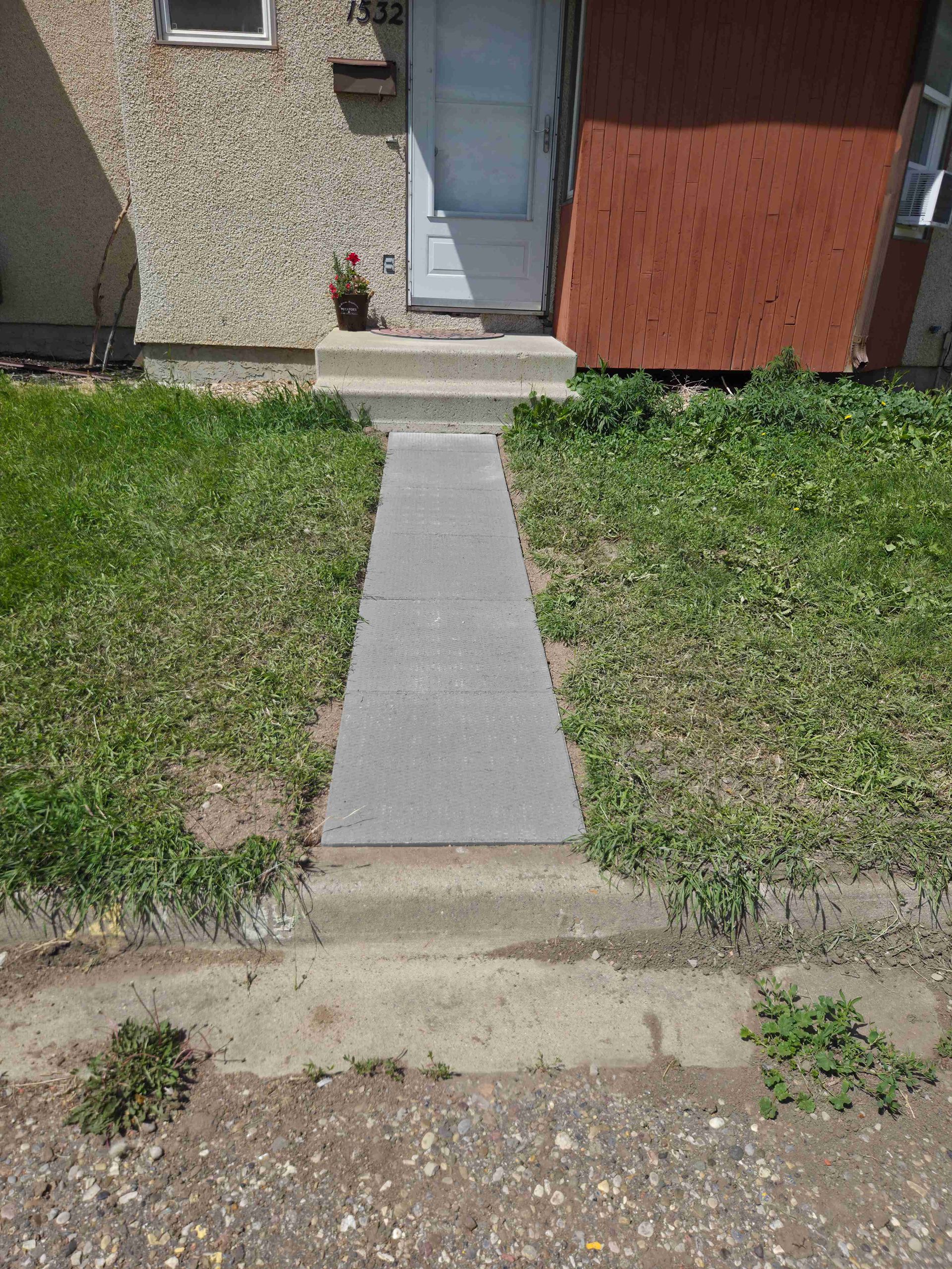 Concrete walkway leading to a house entrance, bordered by grass and gravel.