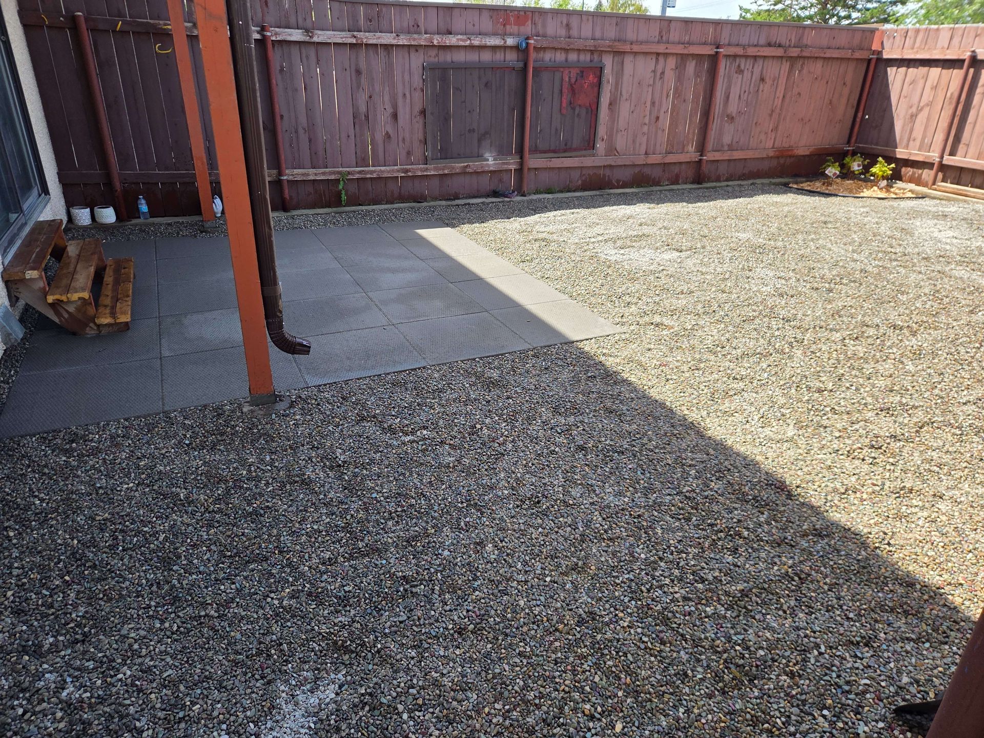 Gravel and concrete backyard patio beside a wooden fence, with a small covered area and metal post.