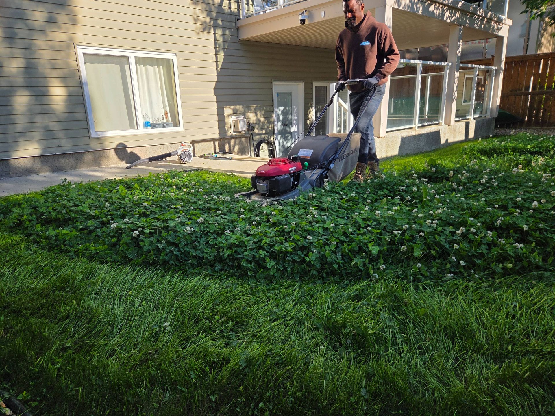 Person mowing a lush green lawn beside a beige apartment building with a red push mower.