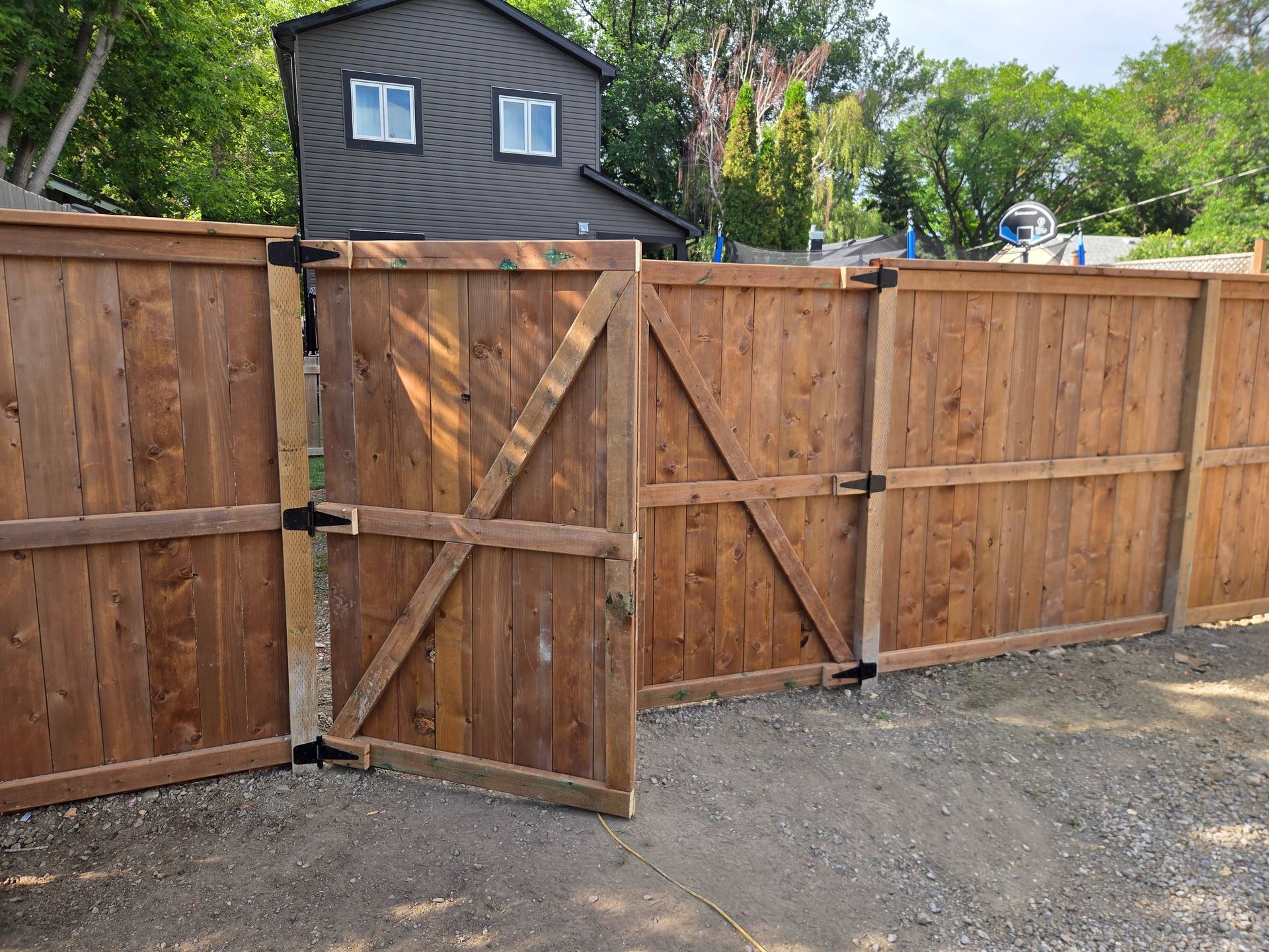 Wooden backyard fence with an open gate, gravel ground, and a dark shed visible behind it.