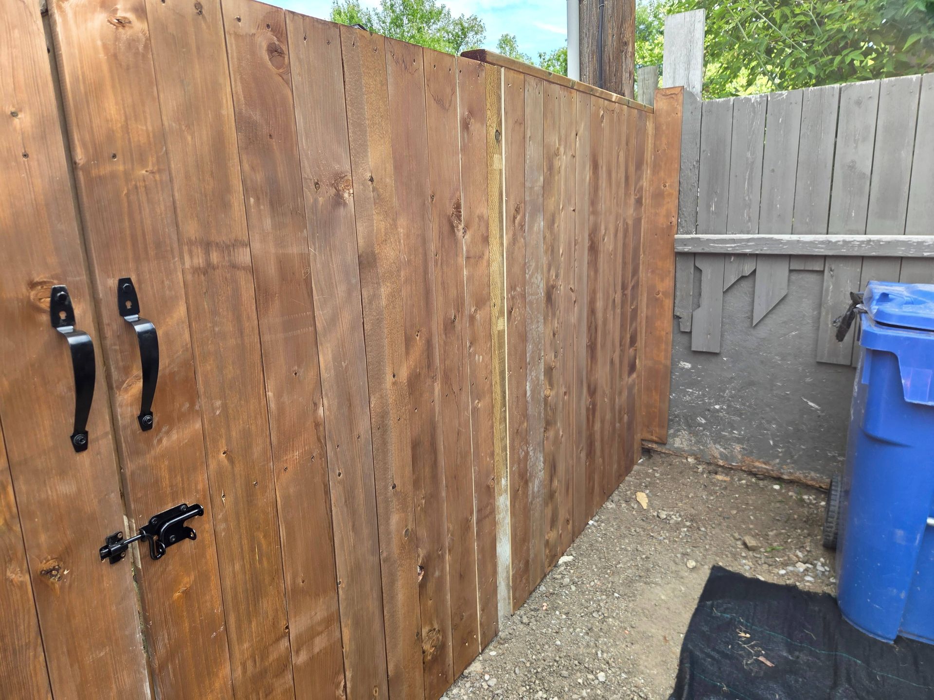 Wooden fence corner beside a gray gate and blue recycling bin on a paved side yard.