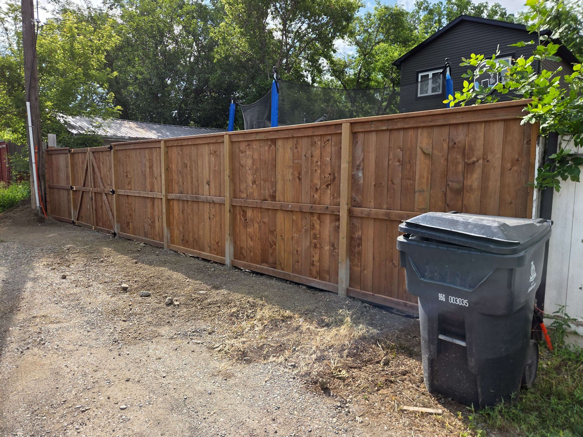 Wooden privacy fence along a gravel driveway, with a black trash bin in front and a house behind trees.