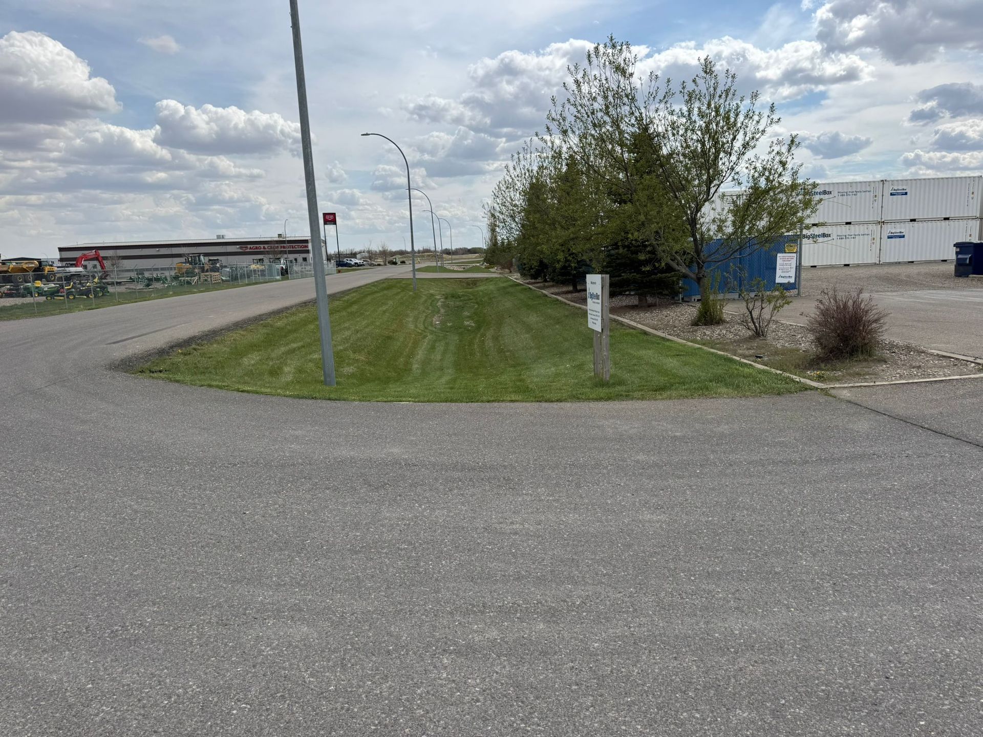Grassy median beside a road with a signpost, trees, and industrial buildings under a cloudy sky