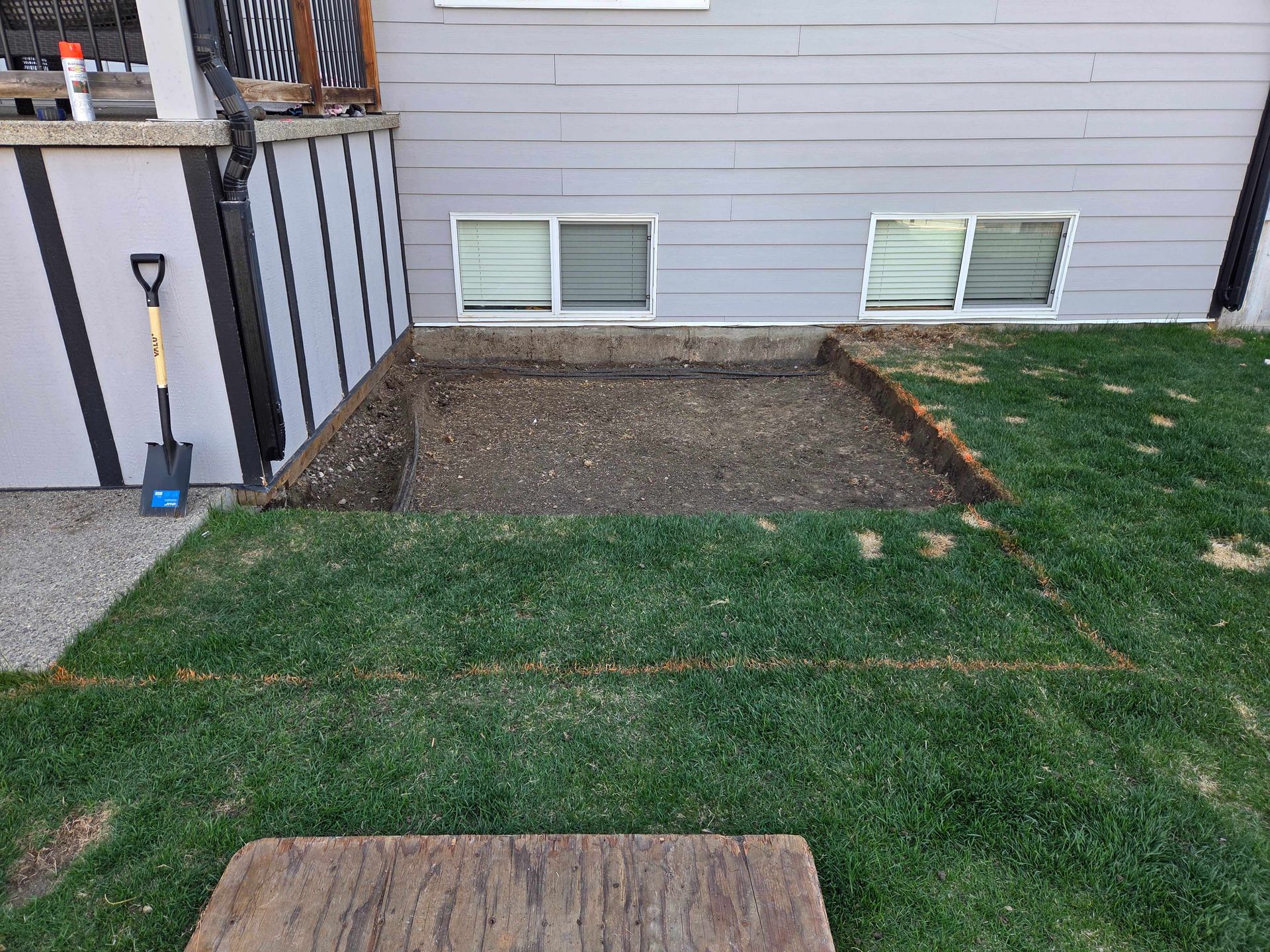 Backyard with a bare dirt patch beside a house, bordered by green lawn and basement windows.