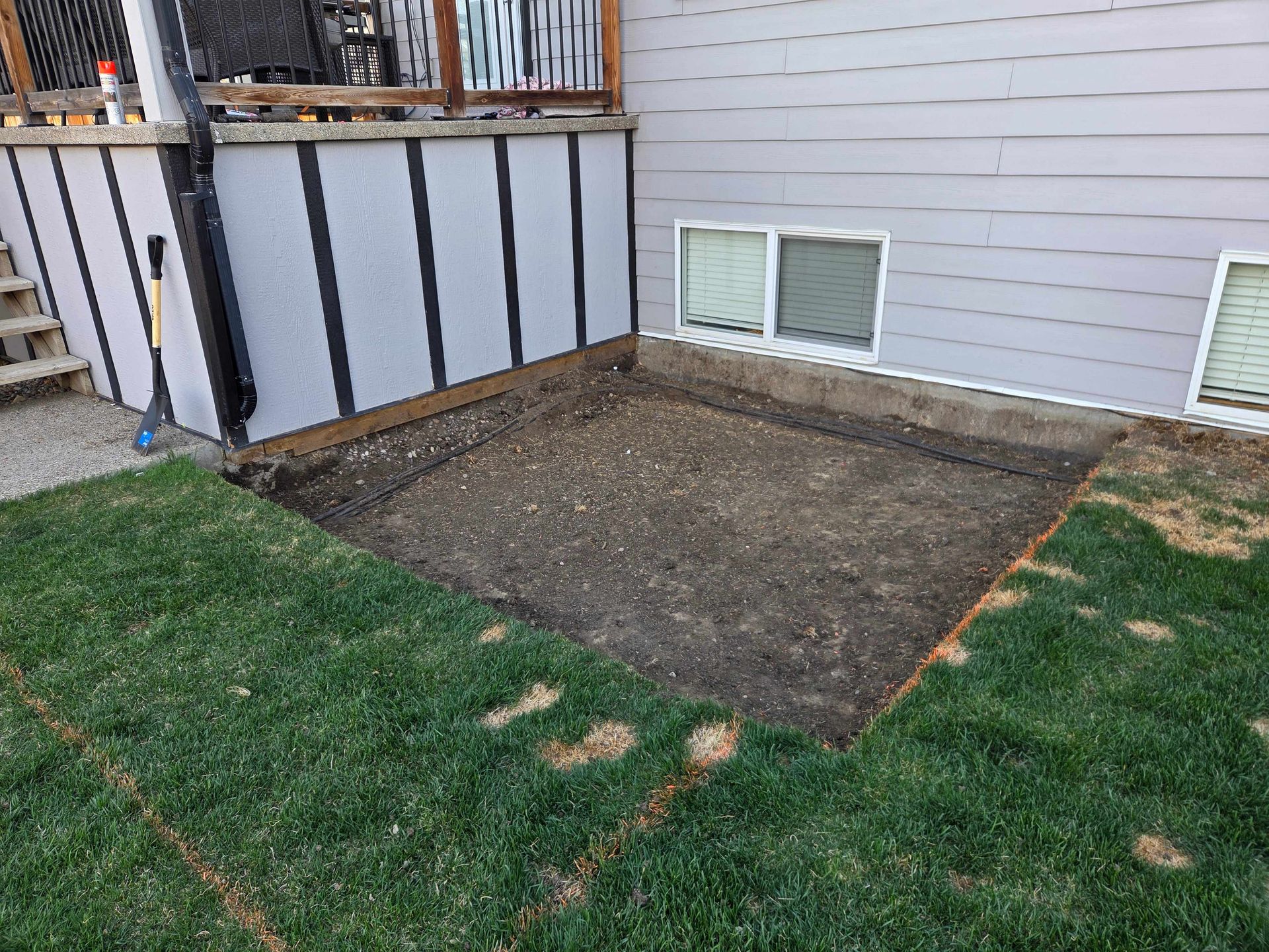 Backyard corner with a rectangular bare dirt patch beside a house and green grass nearby
