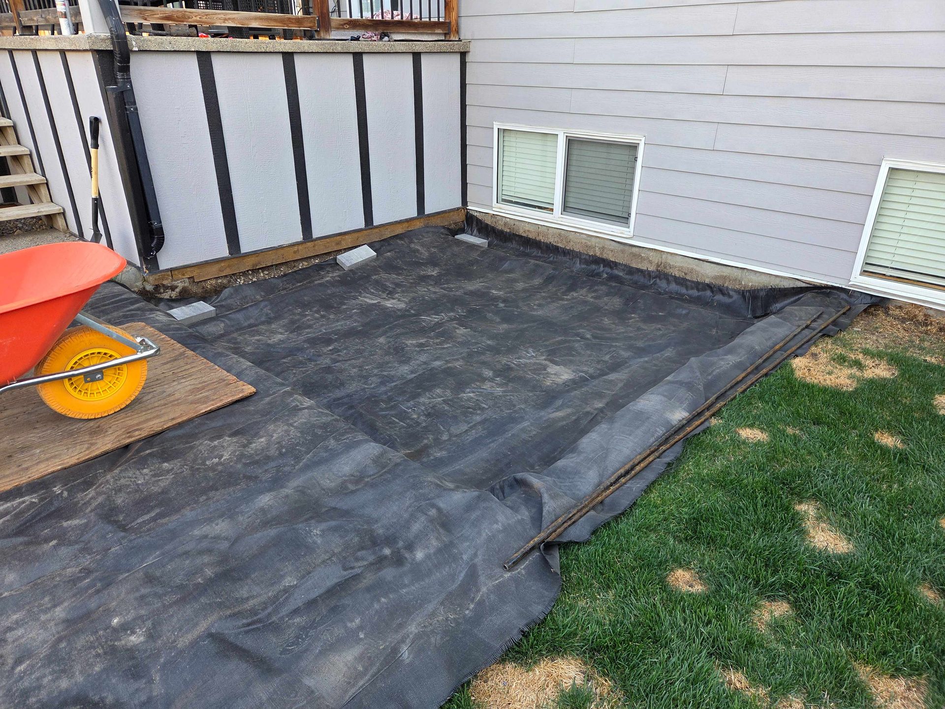 Backyard area with black weed barrier, house siding, basement windows, and a wheelbarrow on the left