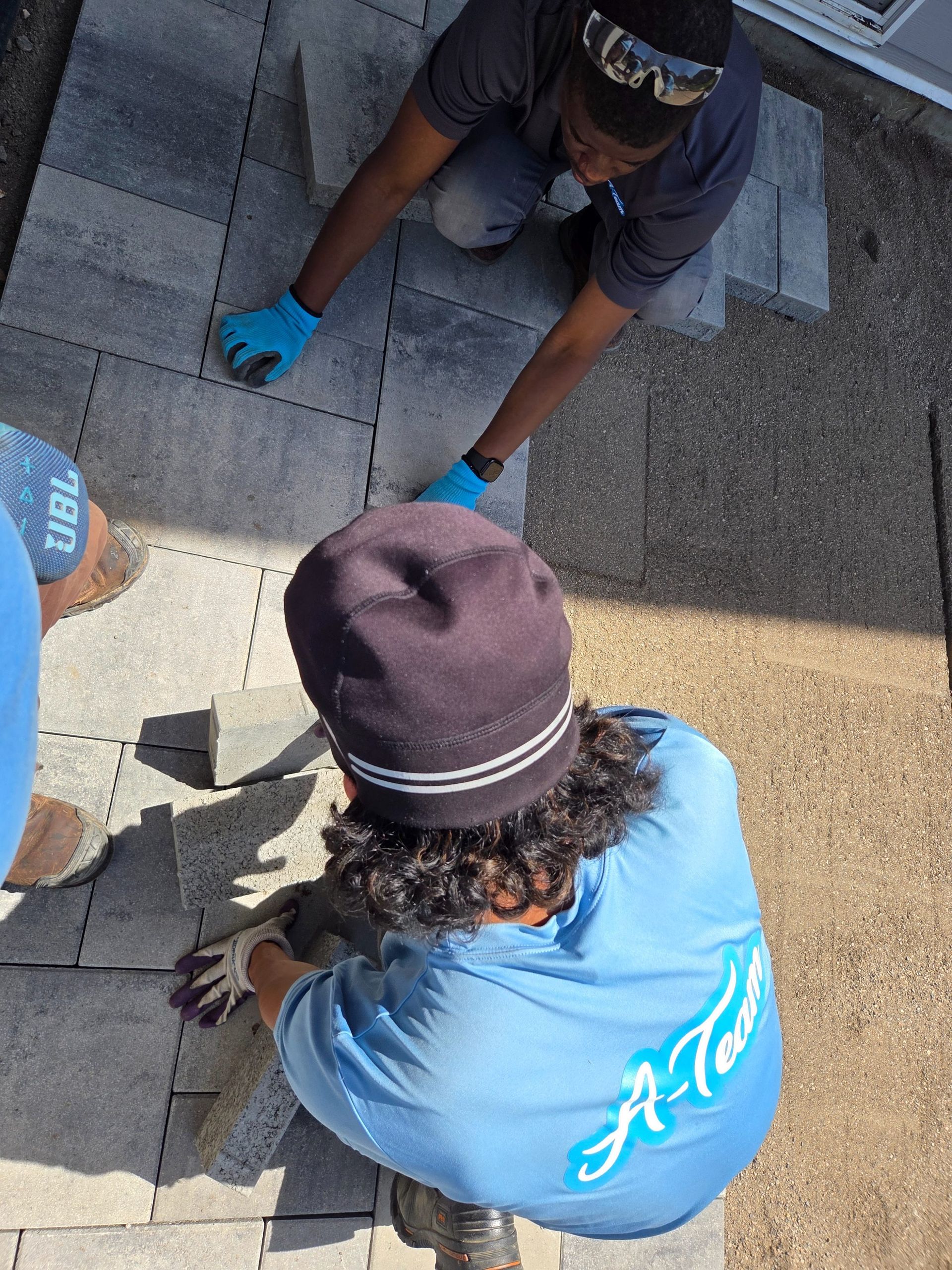 Two workers in blue gloves kneel on a rooftop, handling shingles near a roof repair area.