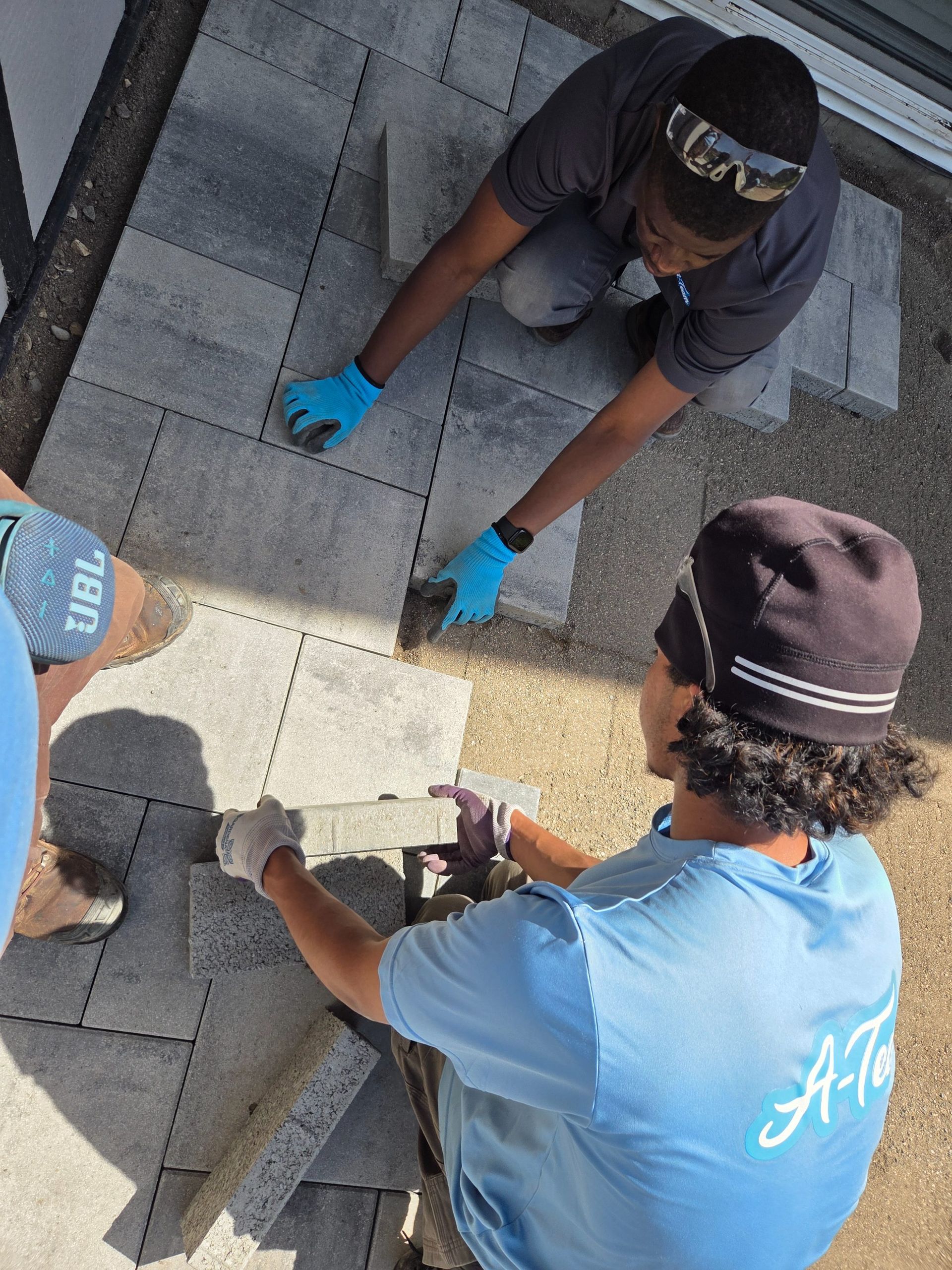 Two workers in blue gloves and shirts install gray roof shingles with hand tools.