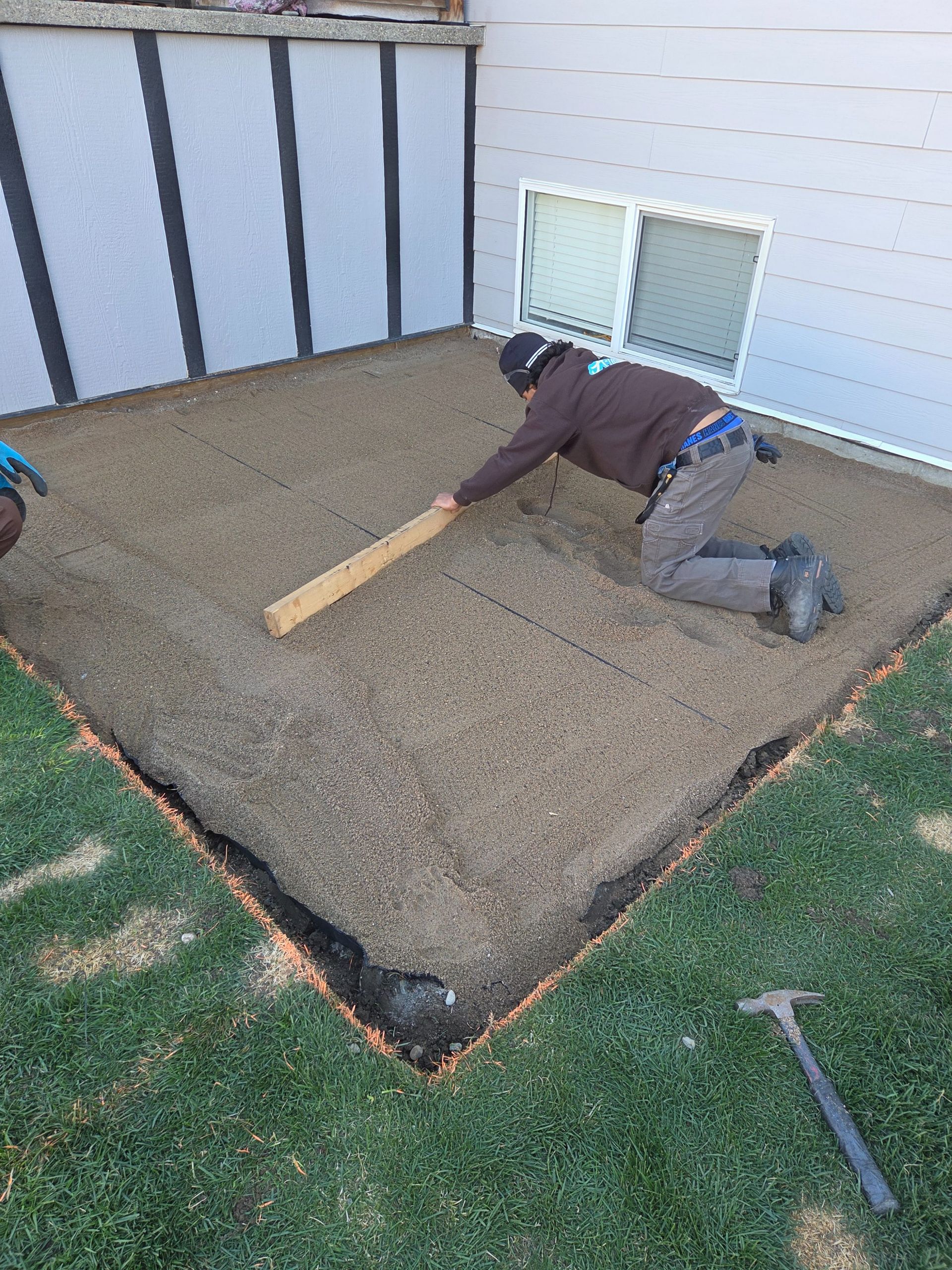 Worker smoothing gravel in a backyard patio area beside a house.