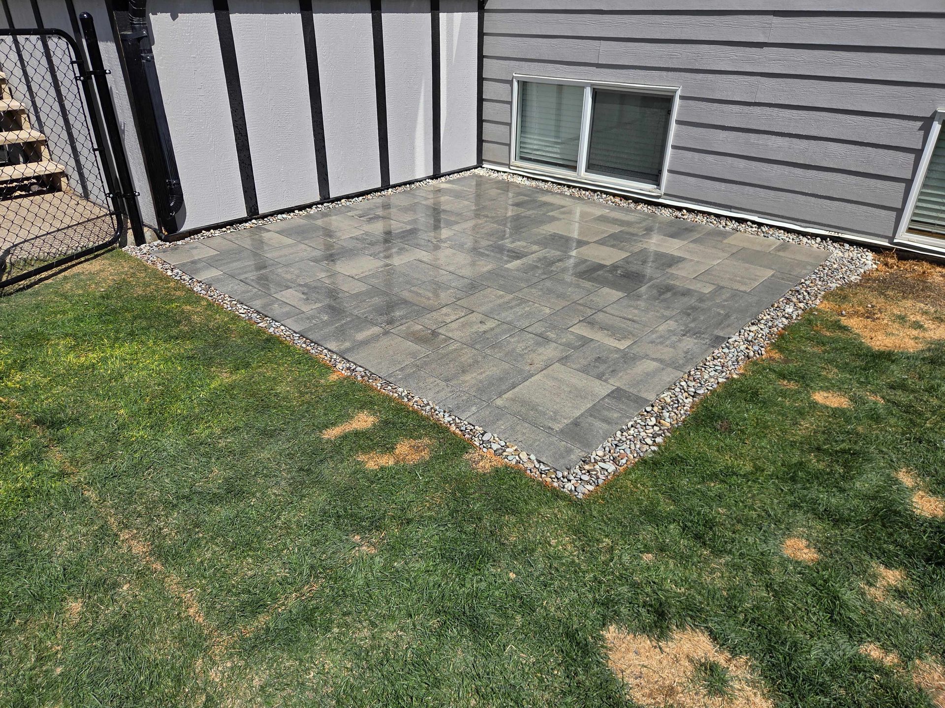 Small gray stone patio beside a house, bordered by grass and gravel, with a basement window and exterior stairs nearby