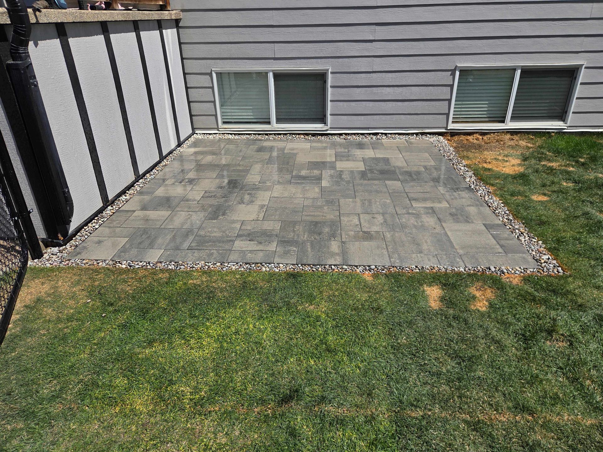 Paver patio beside a house with windows, bordered by grass and a stone edge