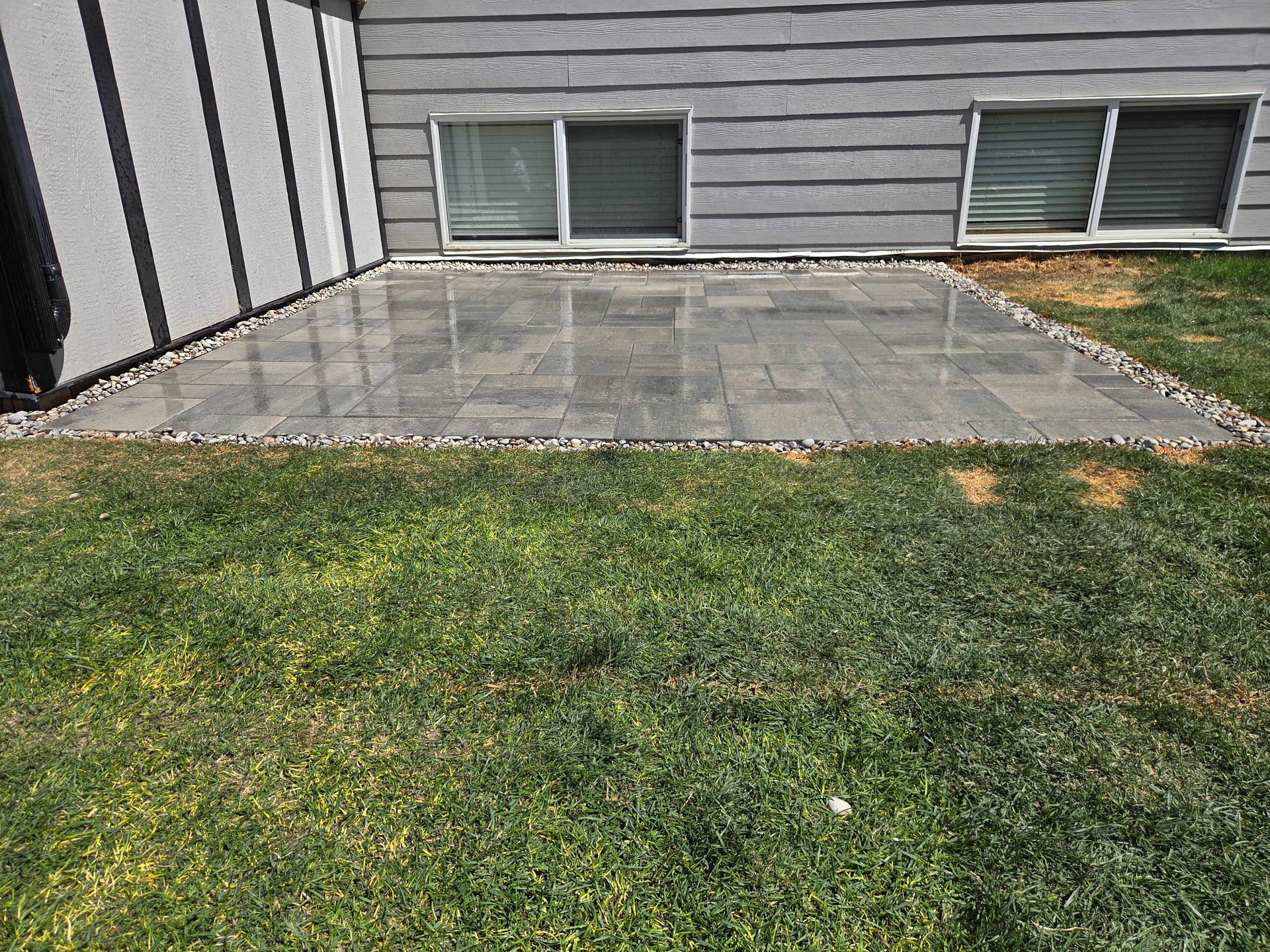 Backyard patio with gray pavers beside a house, bordered by grass and windows.