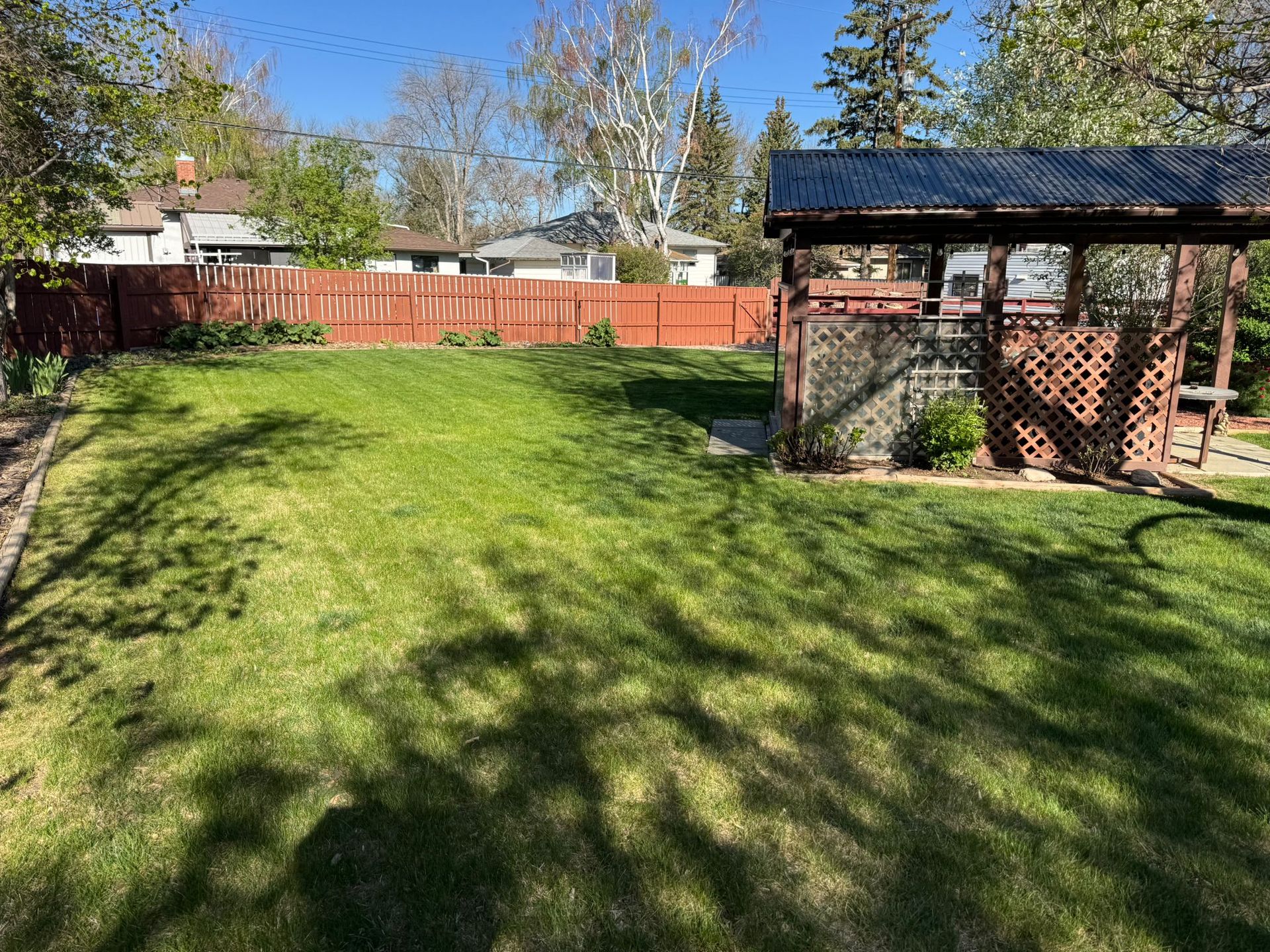 Sunny backyard with green lawn, wooden fence, and a brick gazebo under trees