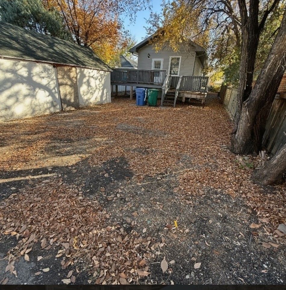 Leaf-covered backyard with a small house, garage, trash bins, and trees in autumn.