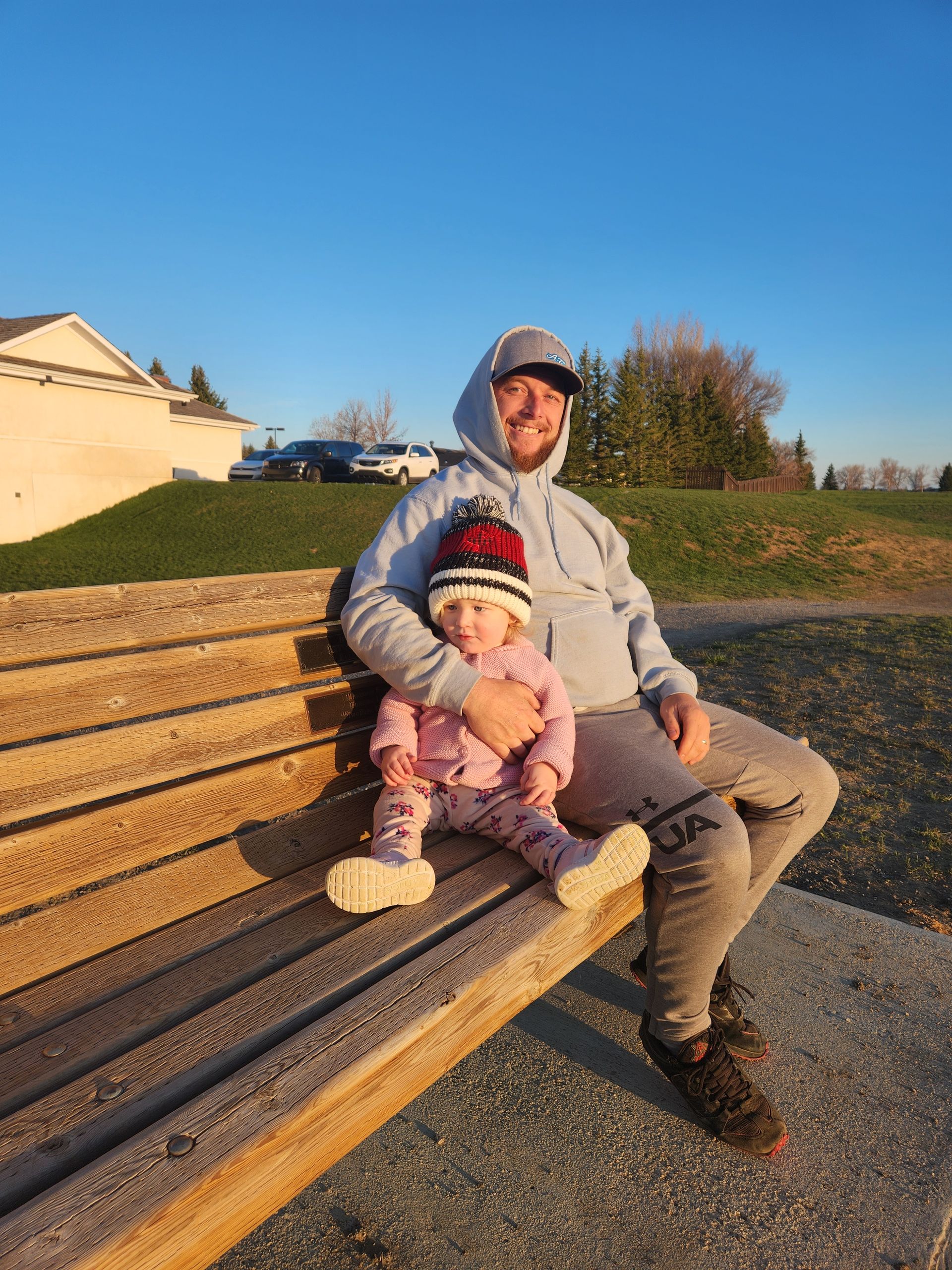 Man and child sitting on a wooden bench in a sunny park, both wearing winter hats and jackets.