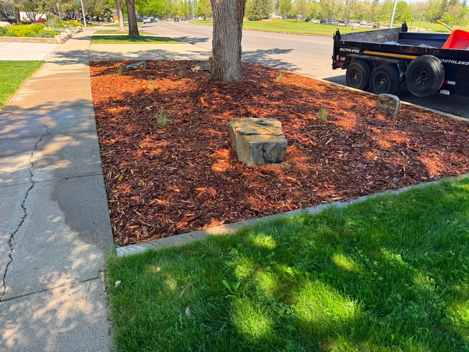 Mulched tree bed beside a sidewalk and grassy lawn, with a stone marker and a trailer parked nearby.