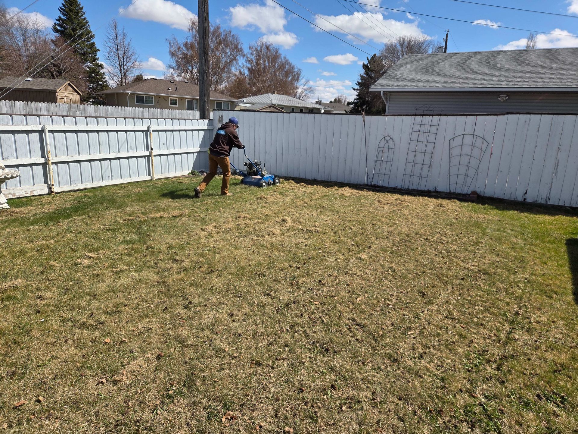 Person using a leaf blower in a backyard beside a white fence and shed on a sunny day
