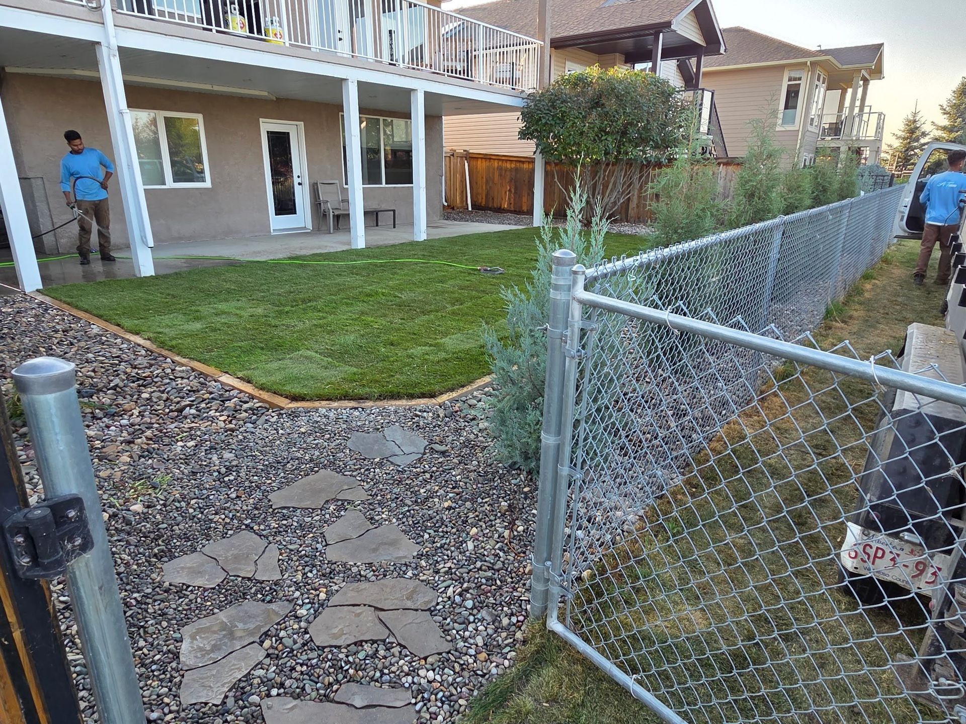 Backyard with stone path, grassy lawn, chain-link fence, and two people near a house.