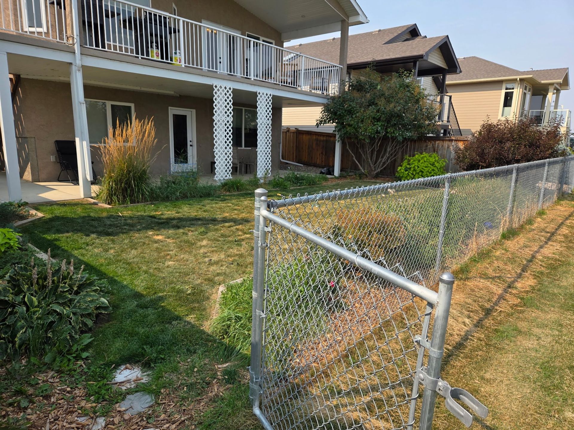 Suburban backyard with chain-link fence, green lawn, and two-story house with balcony and patio