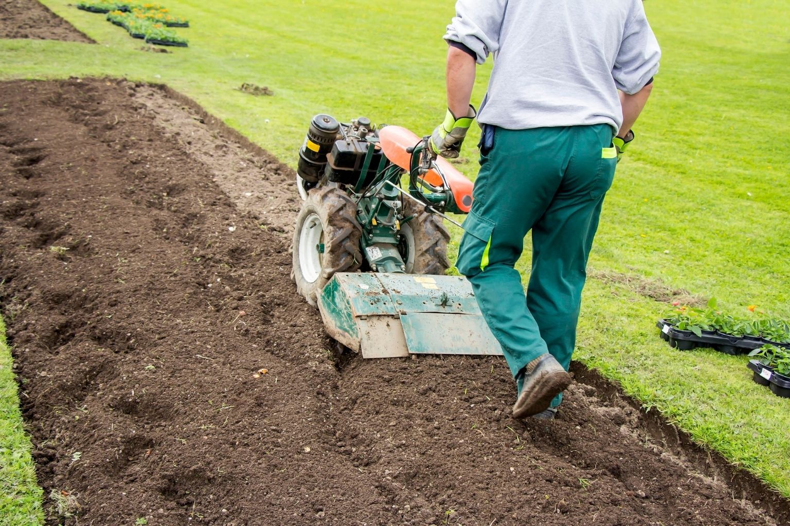 A person operates a motorized tiller to cultivate soil in a garden bed.