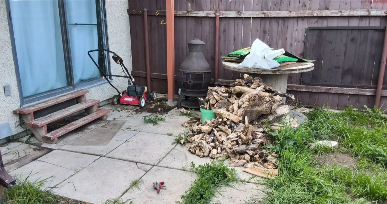 Backyard patio with pavers, a wooden fence, a grill, and a pile of chopped wood and yard debris