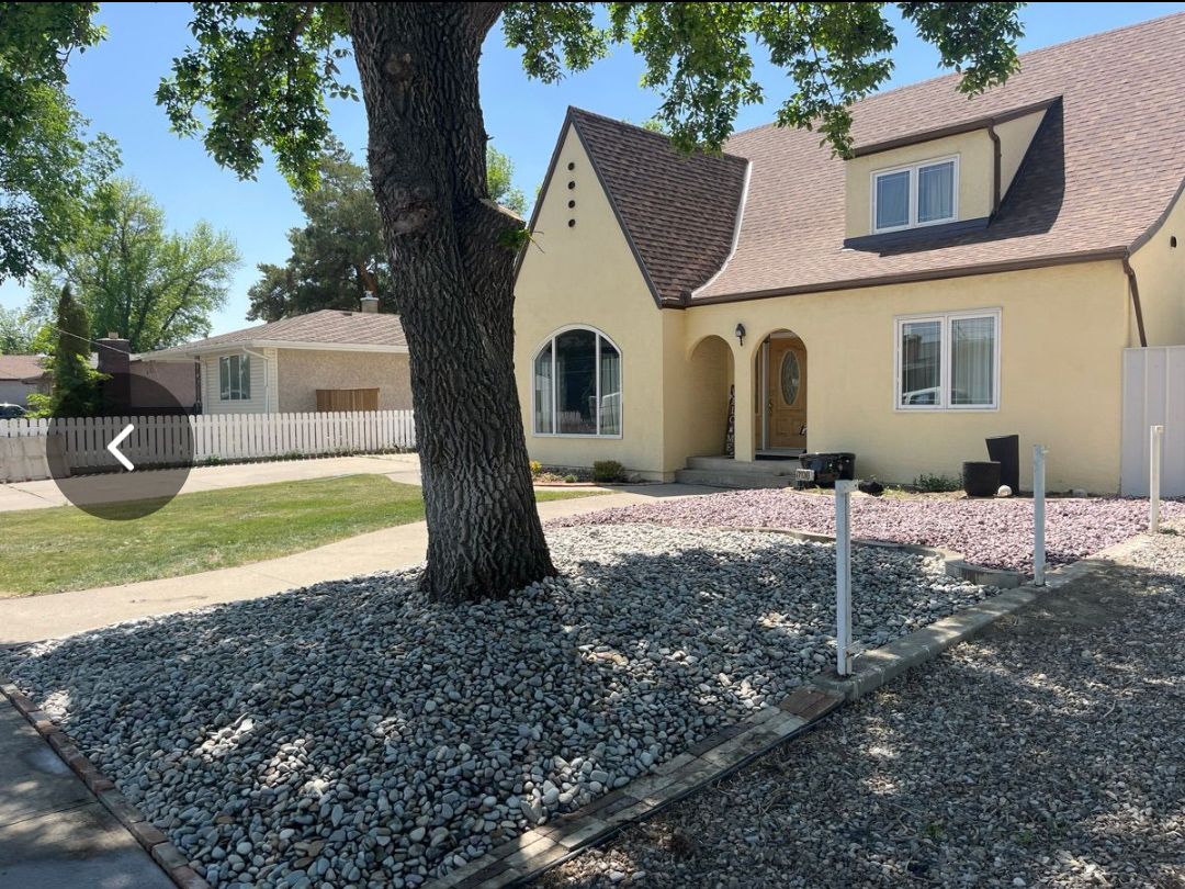 Suburban house with tan stucco, brown roof, front yard rocks, and a large tree in the foreground