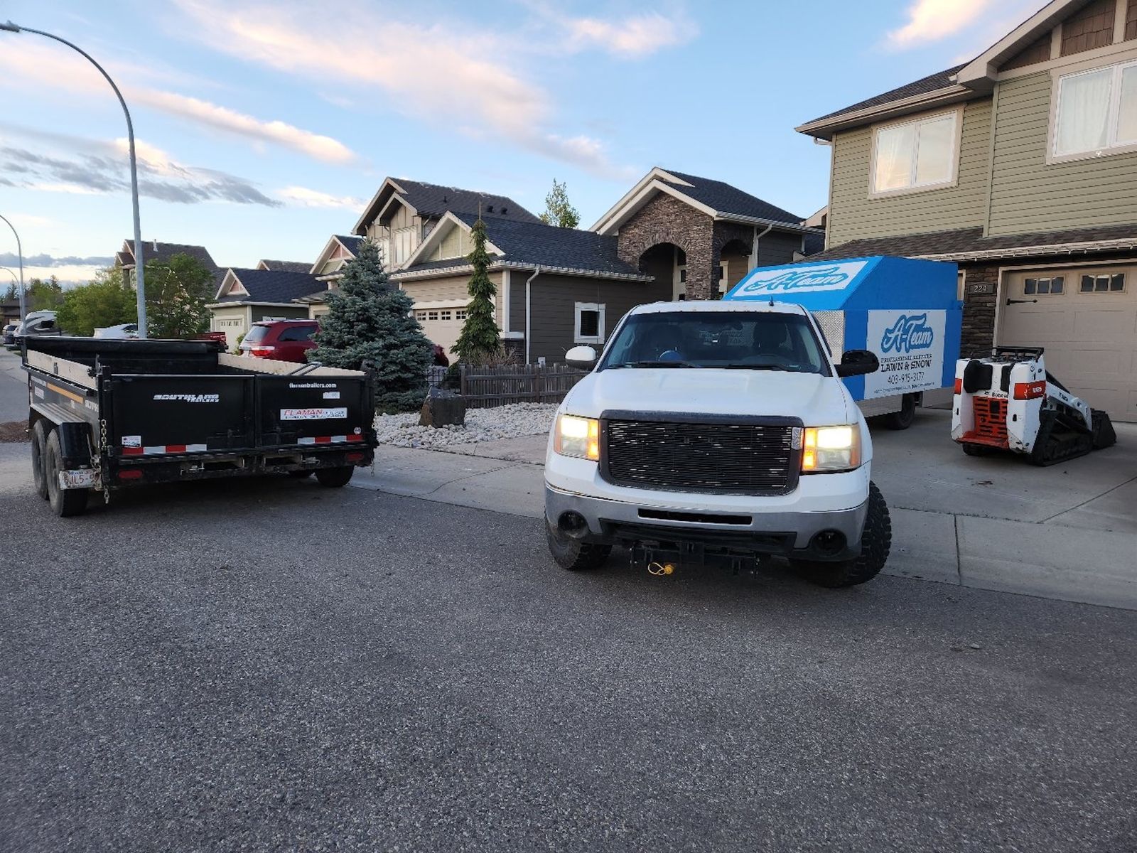 A white pickup truck parked in a driveway next to a trailer and equipment, with another trailer parked on the street.
