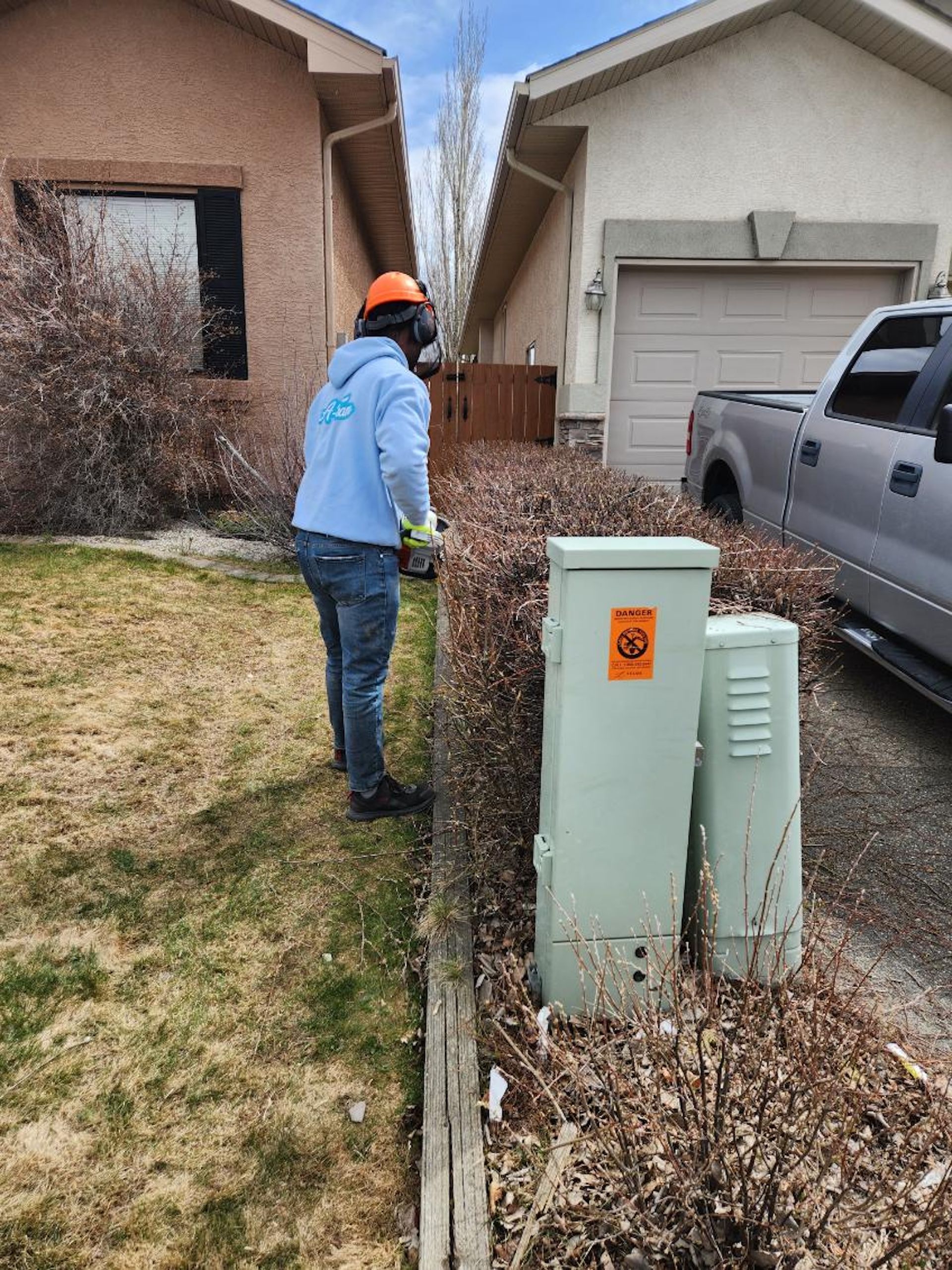A person wearing a hard hat and light blue hoodie trims a hedge near two utility boxes beside a residential driveway.
