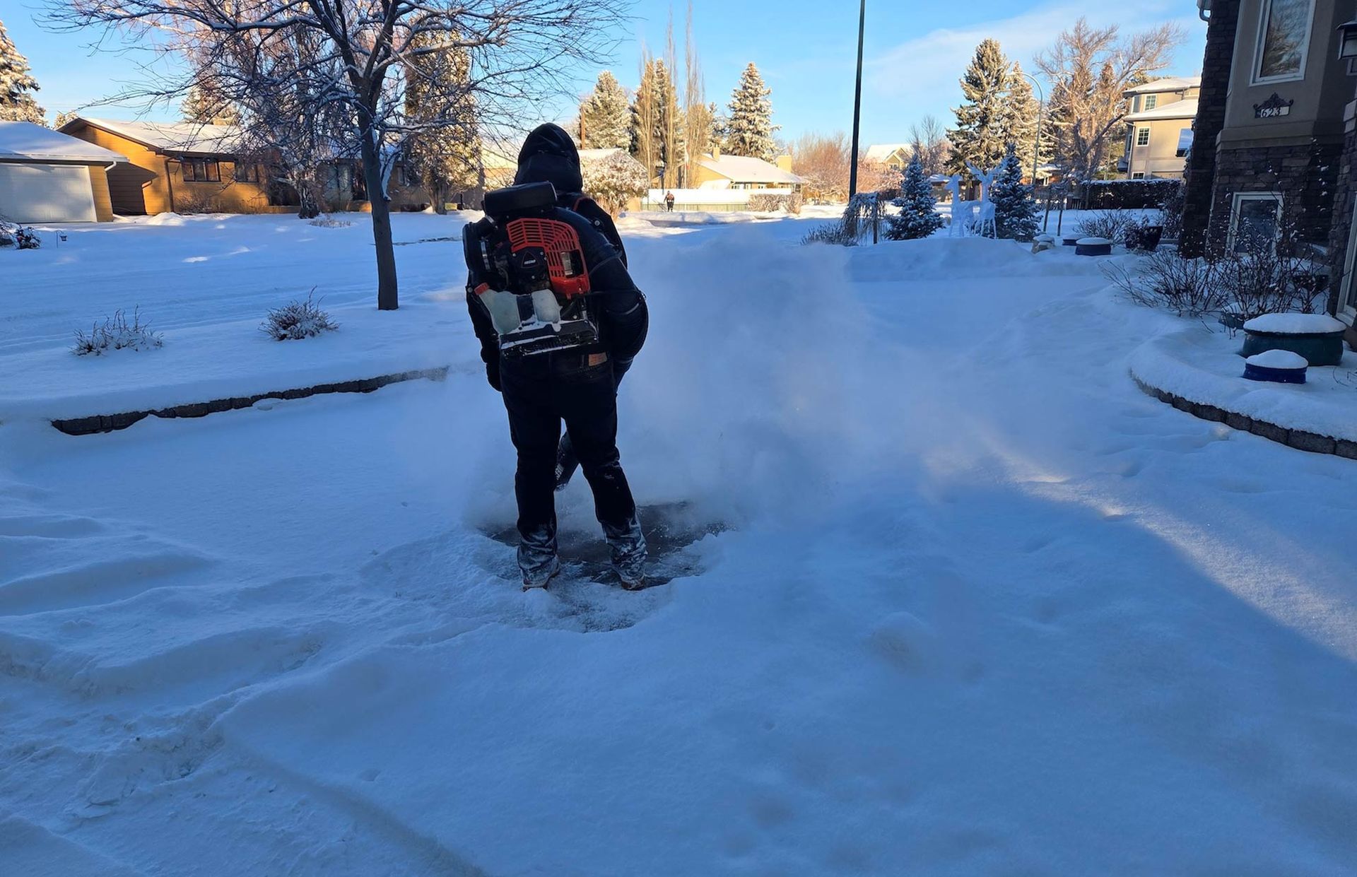 A person using a backpack leaf blower to clear light, powdery snow from a residential driveway on a sunny winter day.