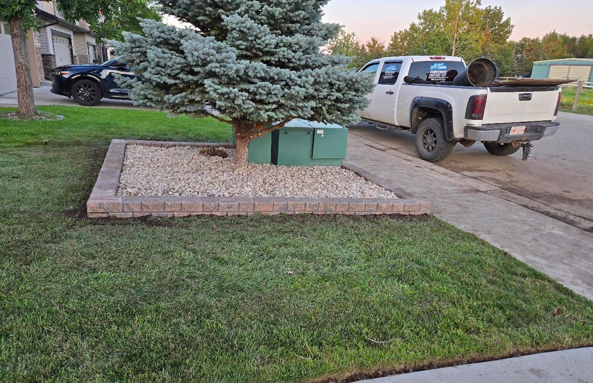 A white pickup truck parked on a residential street next to a landscaped tree bed with gravel and retaining stones.