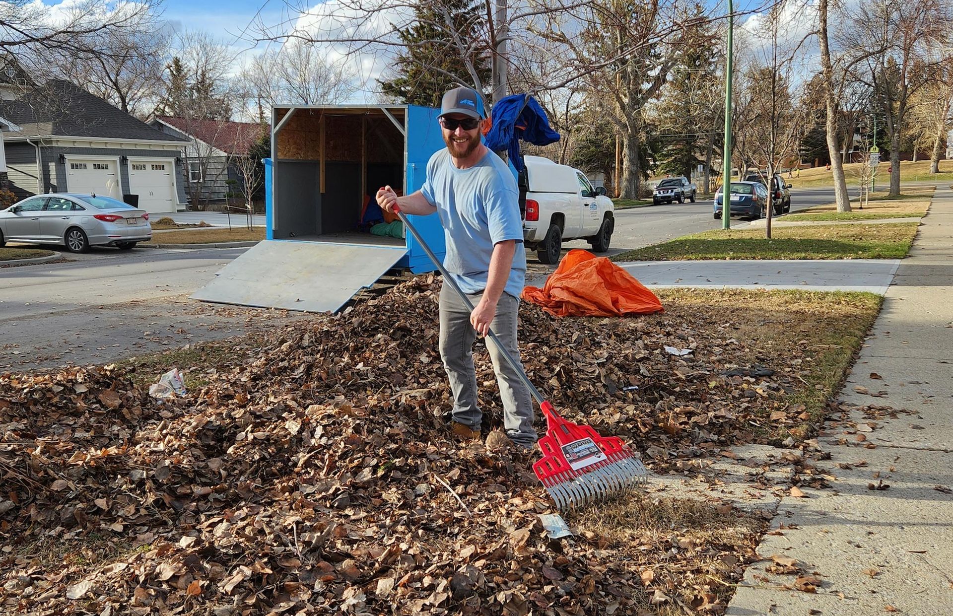 A person in a blue shirt raking brown leaves in a suburban yard with a trailer and parked car in the background.